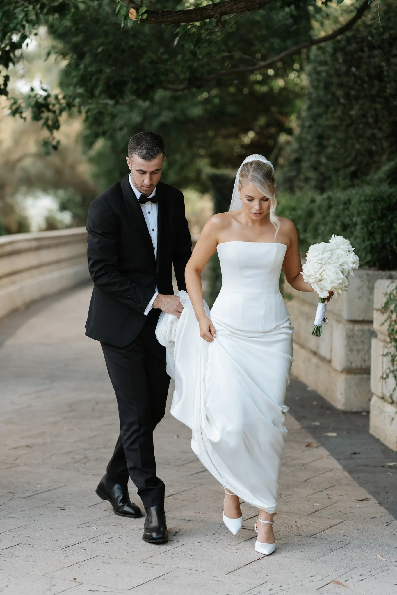 Bride in a white strapless wedding gown holding a bouquet walking with a groom in a black tuxedo on a pathway with greenery.