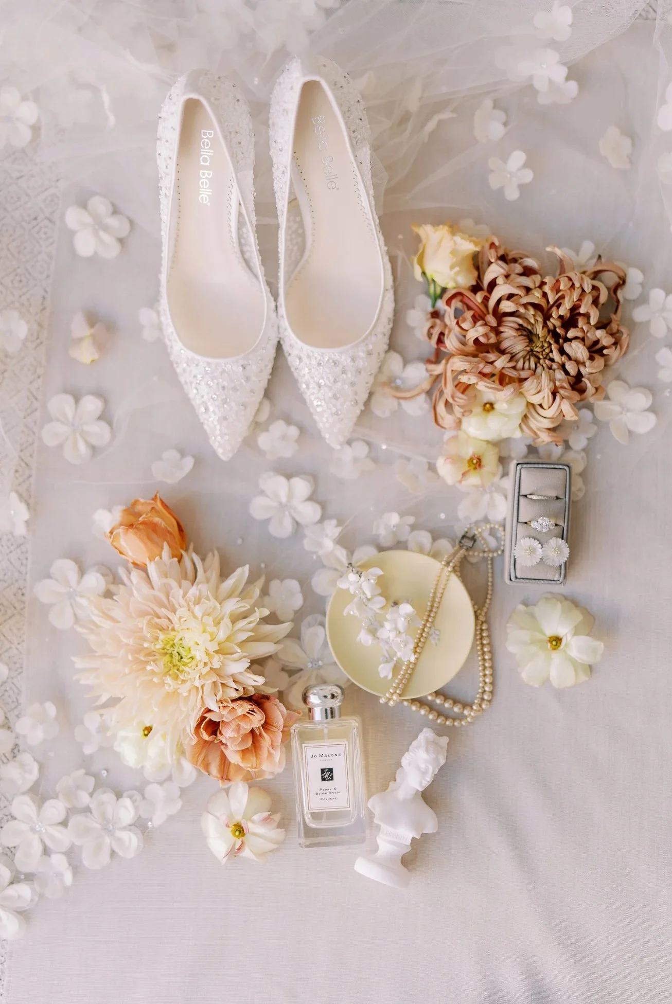 A pair of white high-heeled shoes with embellishments, wedding flowers, and jewelry on a white surface, typically part of a wedding setup.