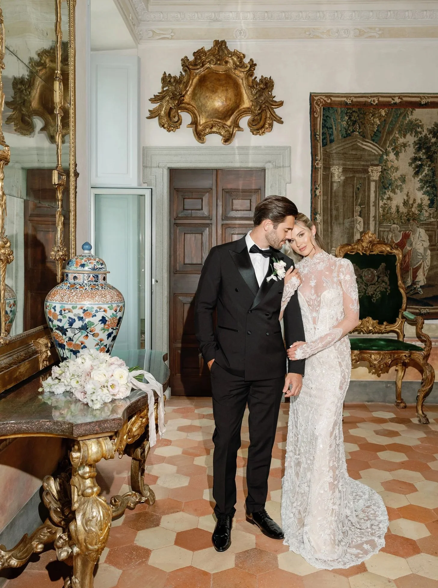 A bride and groom dressed in wedding attire in a historic, ornately decorated room with antique furniture, a large mirror, and artwork on the walls.