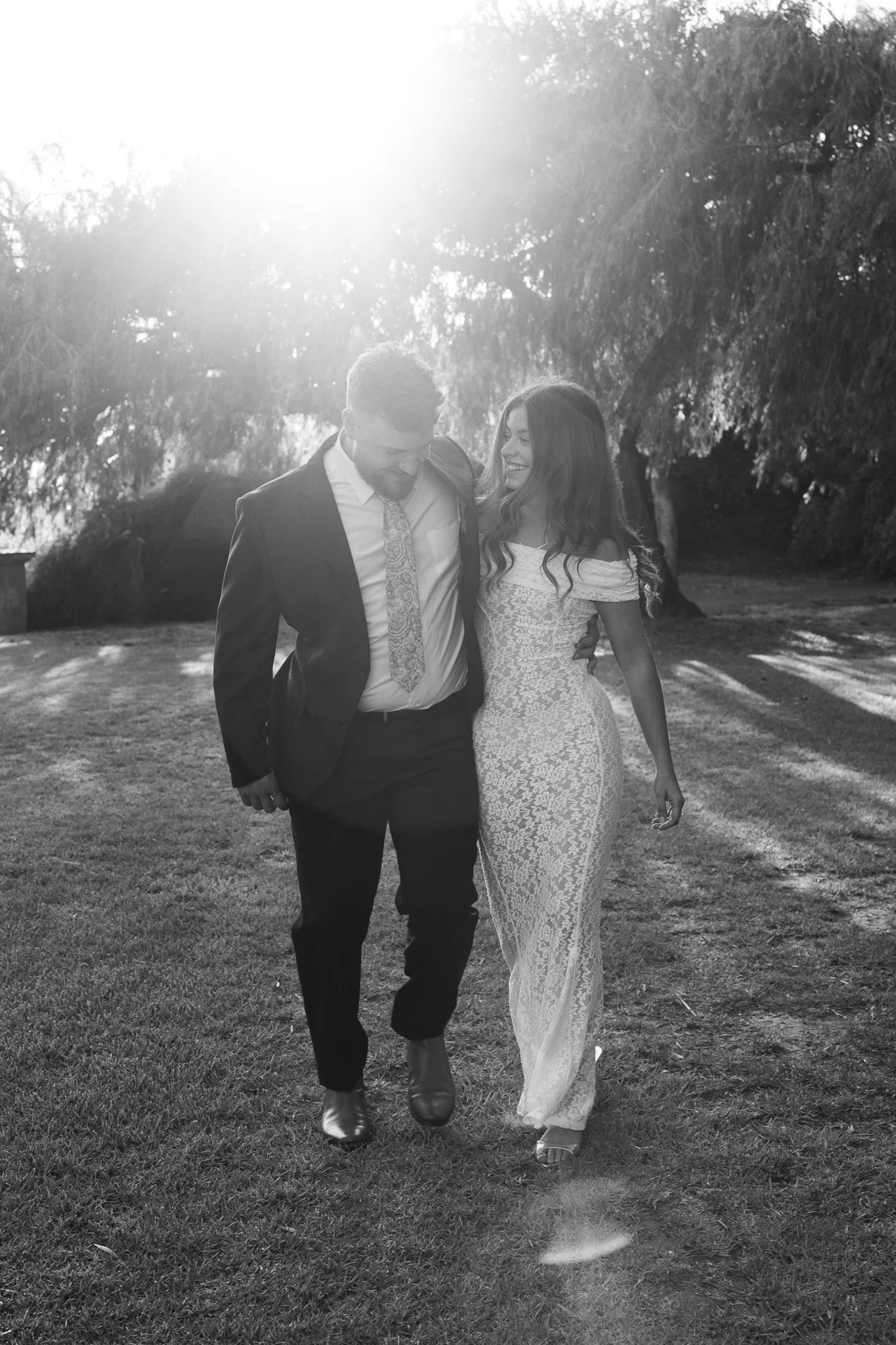 A black and white photo of a couple walking together outdoors, smiling and looking at each other, with sunlight filtering through trees in the background.