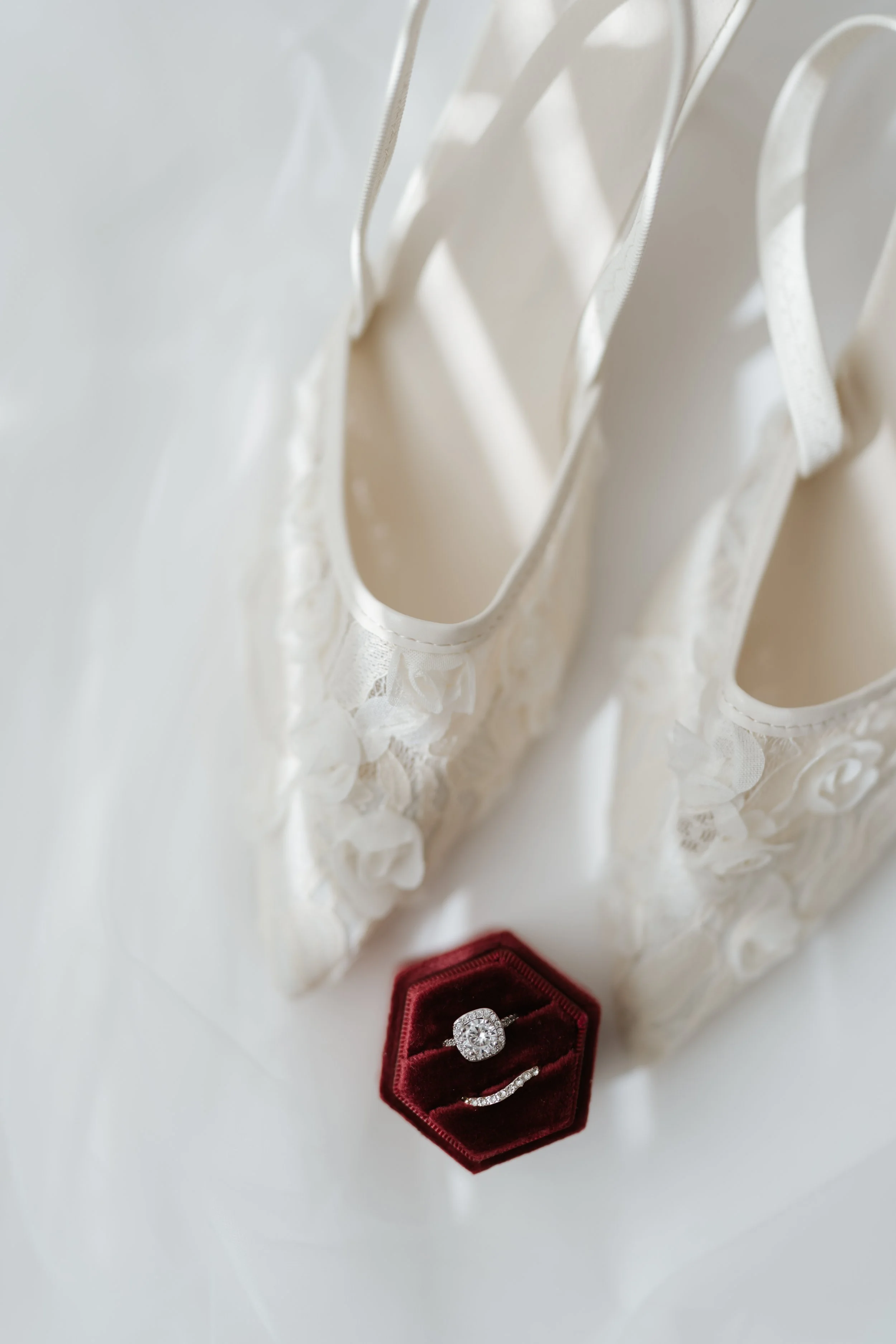 A white wedding dress hanging, with a red velvet ring box containing a diamond engagement ring and wedding band placed below it.
