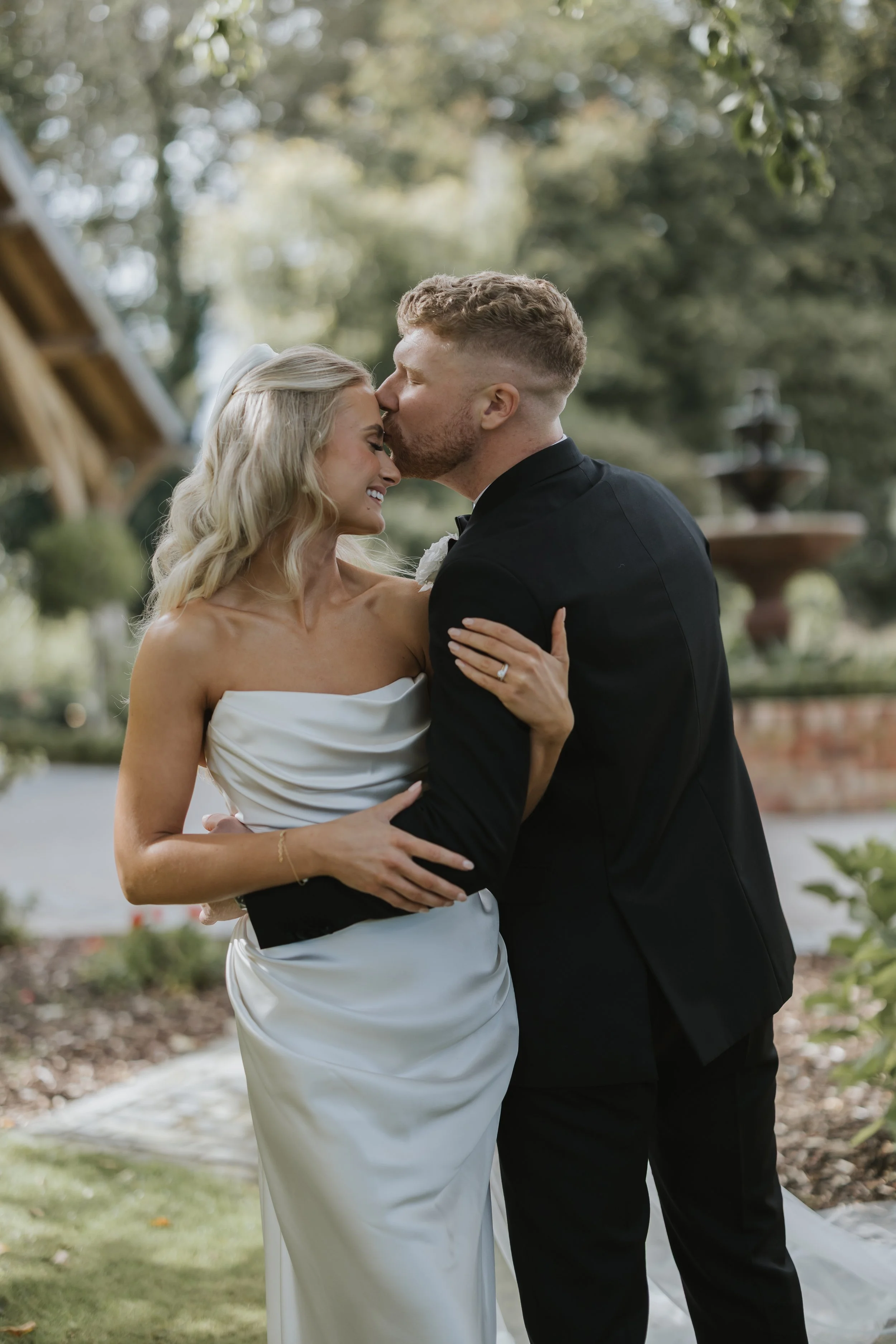 A bride and groom embrace outdoors during their wedding, with the groom kissing the bride on the forehead while she smiles with her eyes closed.