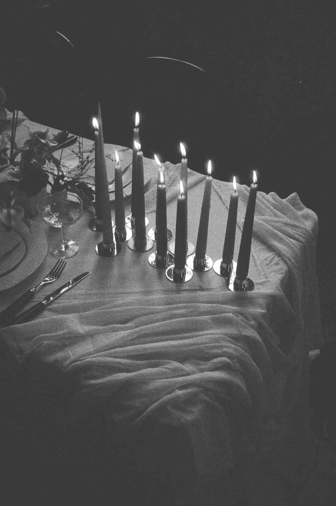 A table with nine lit candles, a plate with cutlery, and a wine glass, covered with a cloth, and decorated with flowers, against a dark background.