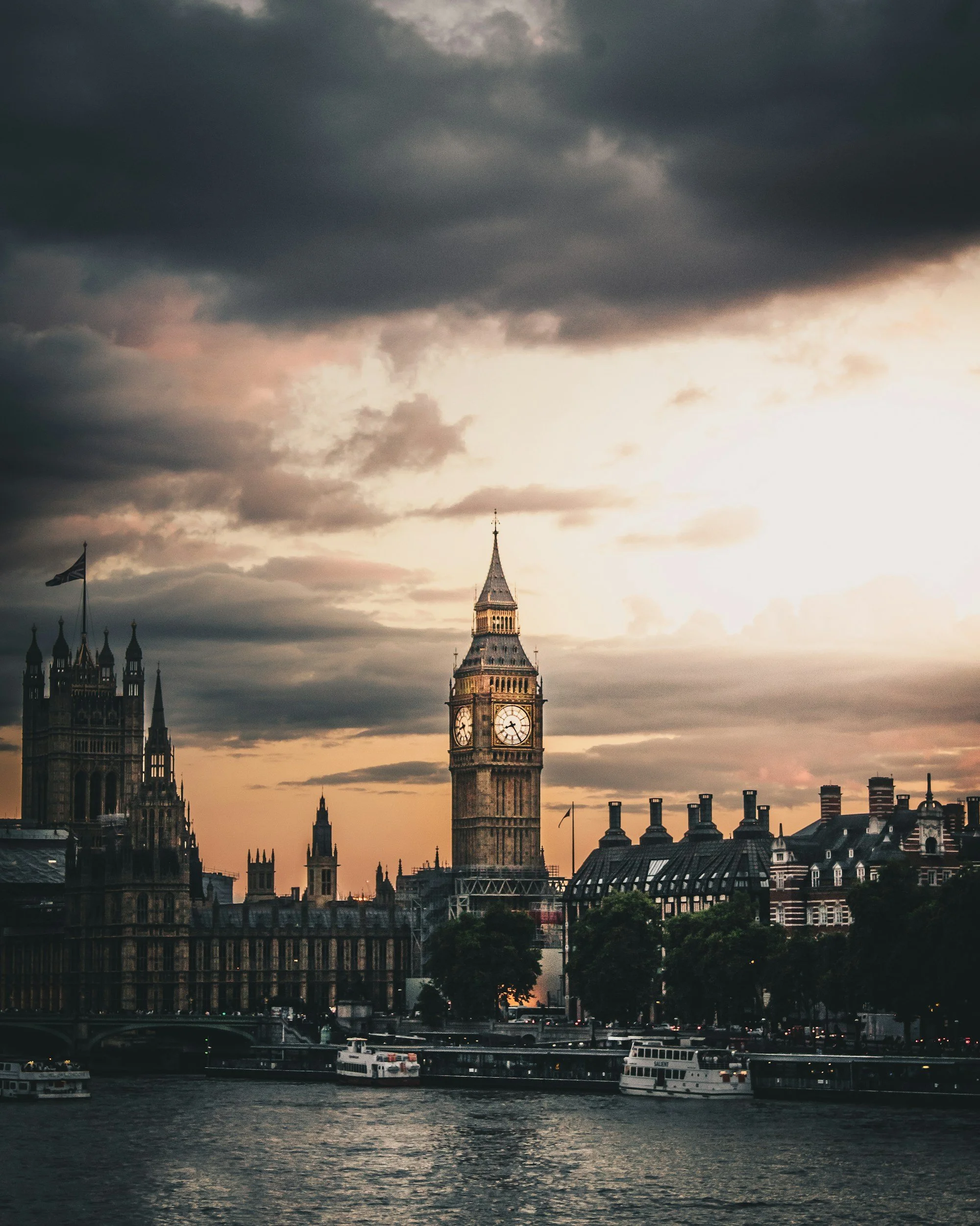 View of the Big Ben clock tower in London with dark clouds overhead and boats on the river in the foreground during dusk or dawn.
