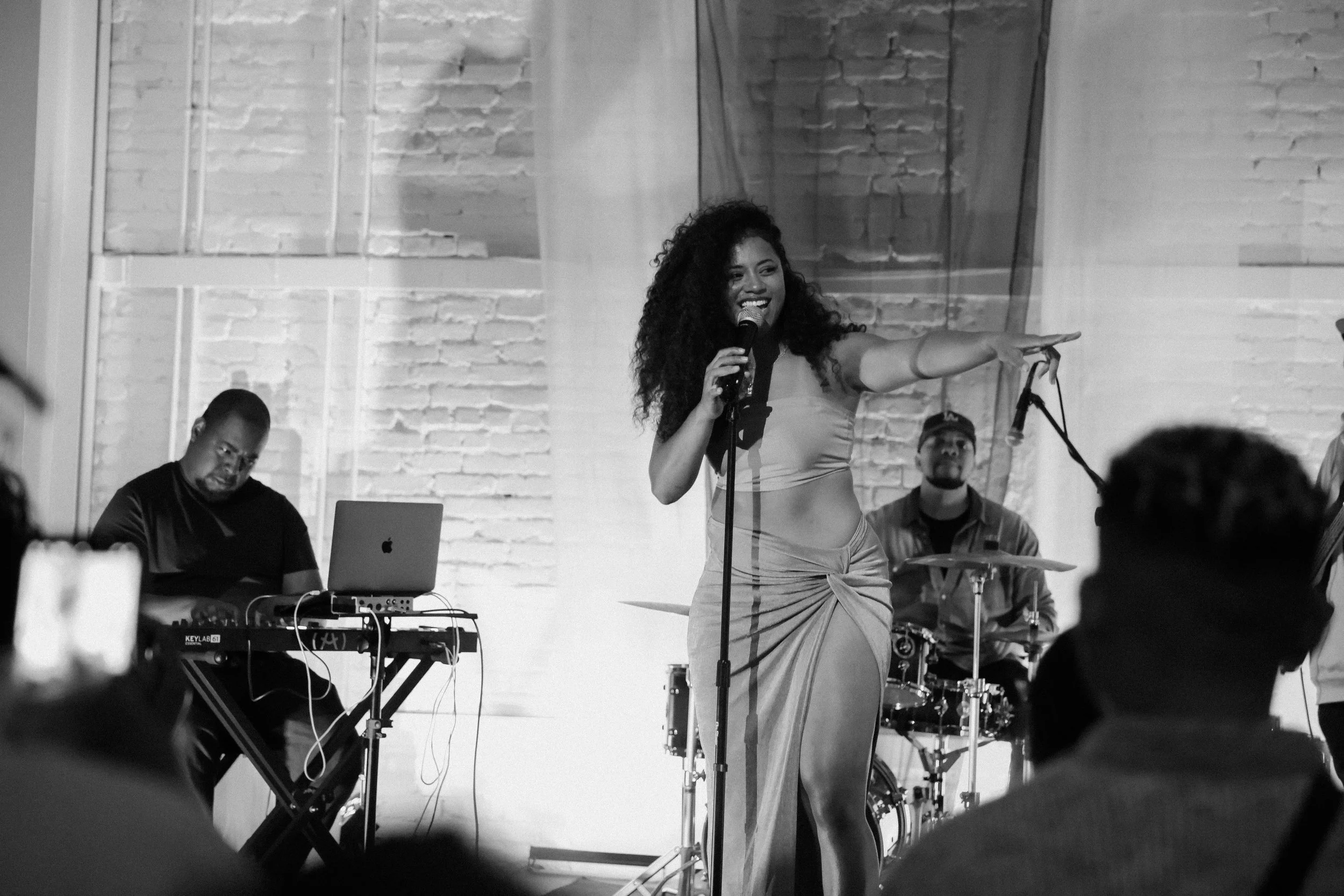 Black and white photo of a female singer with curly hair performing on stage with a microphone, wearing a two-piece outfit and a skirt with a high slit, accompanied by musicians playing drums and keyboard, in a venue with brick walls.