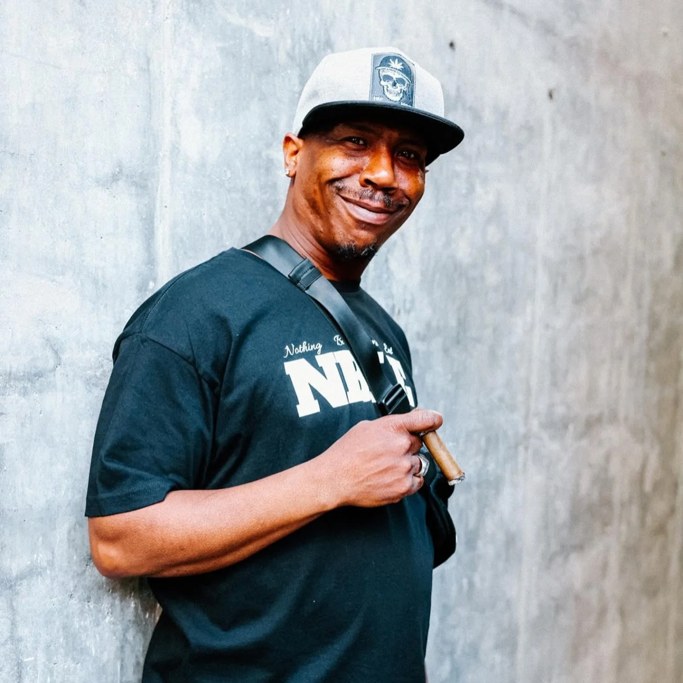 A man with a dark shirt and a gray cap smiling, holding a cigar, standing against a plain gray wall.
