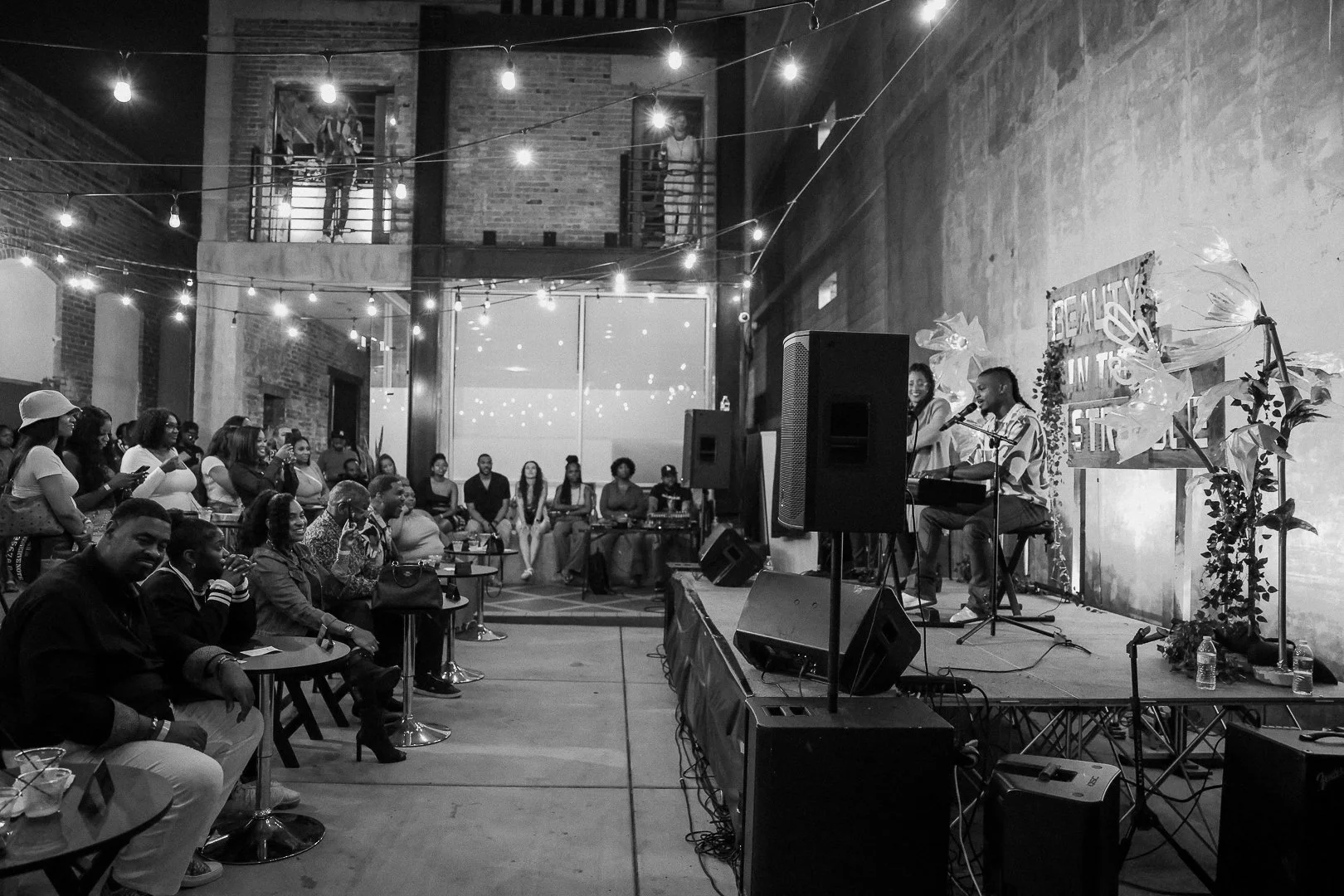 A black and white photo of a live performance in a venue with an audience seated and standing, musical performers on stage with microphones, and string lights hanging from the ceiling.