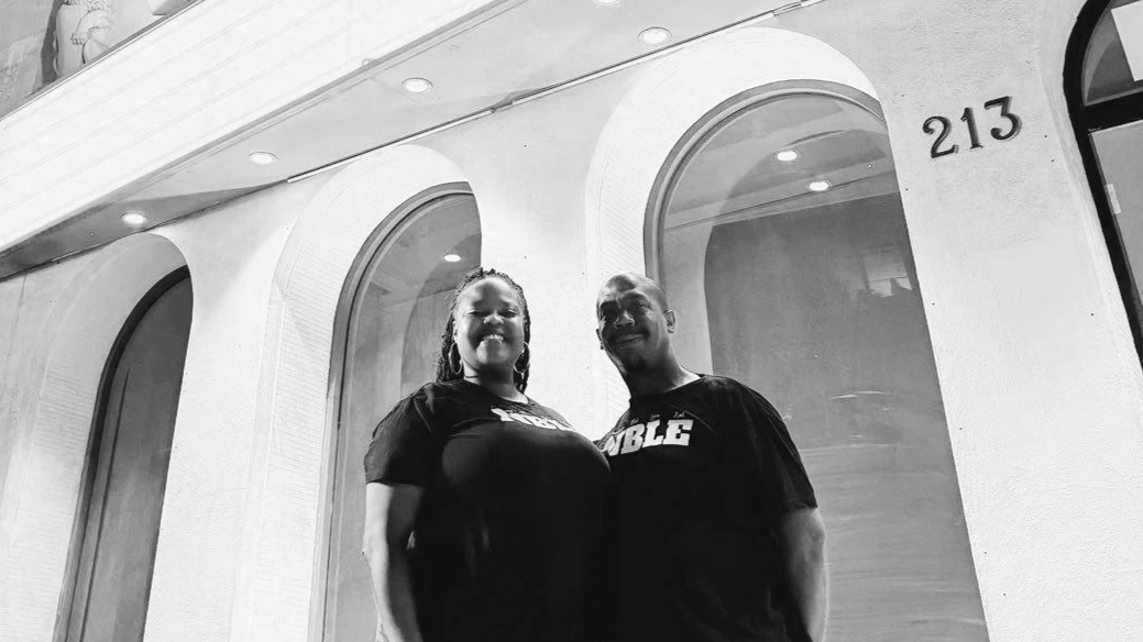 A smiling couple stands outside a building at night, illuminated by exterior lights, with the building's address, 213, visible.