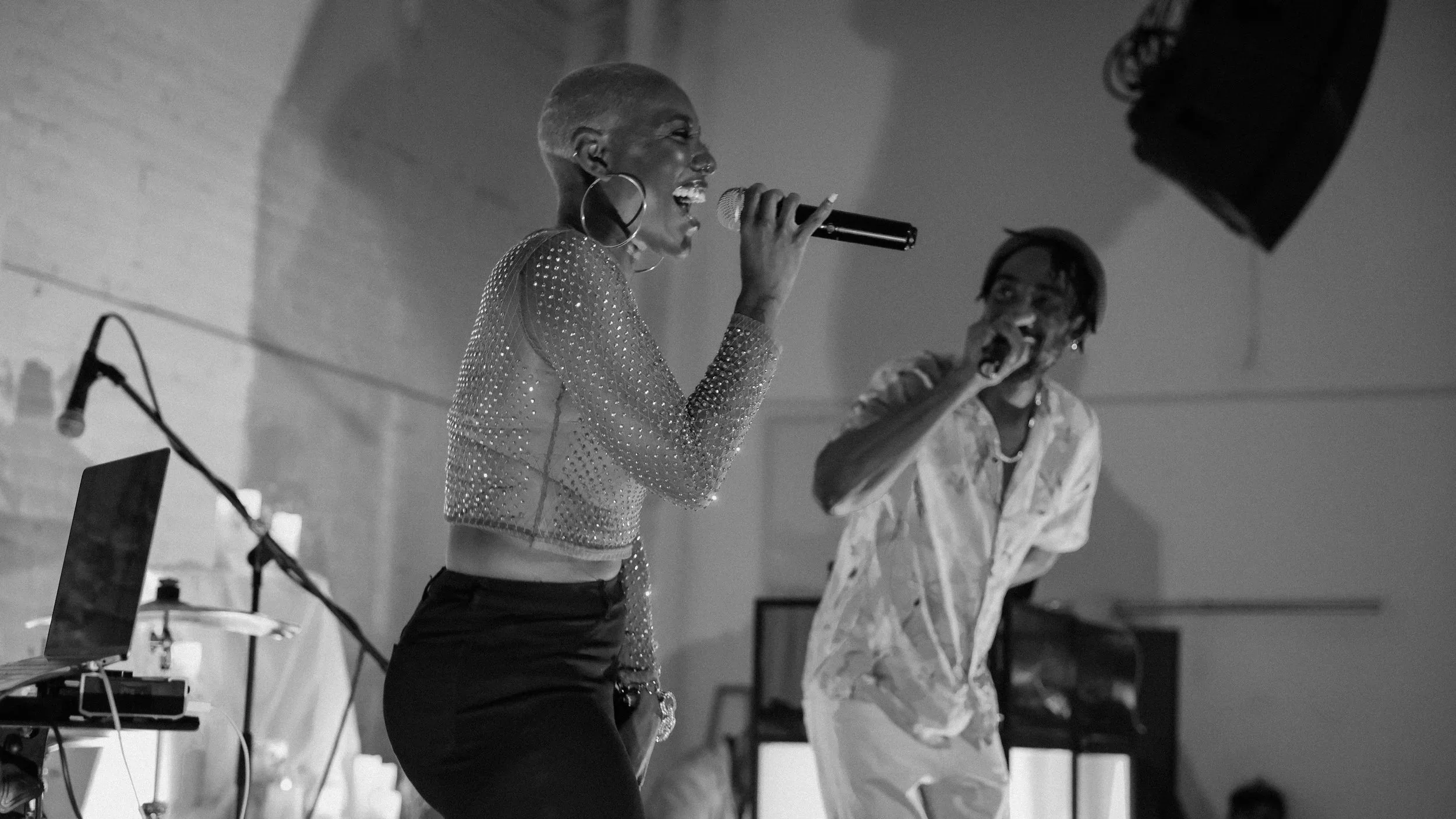 A woman and a man vocalizing into microphones during a performance, with music equipment and speakers visible in the background.