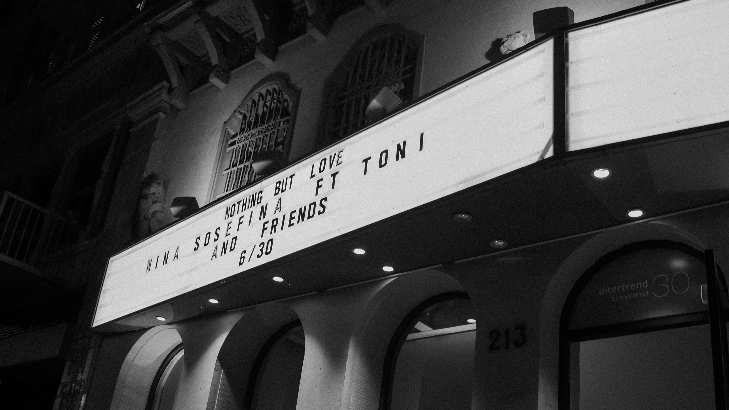 Nighttime photo of a building with illuminated marquee displaying a message about Nina Sosef and love, featuring a mention of Toni and friends, with the date 6/30, in black and white.