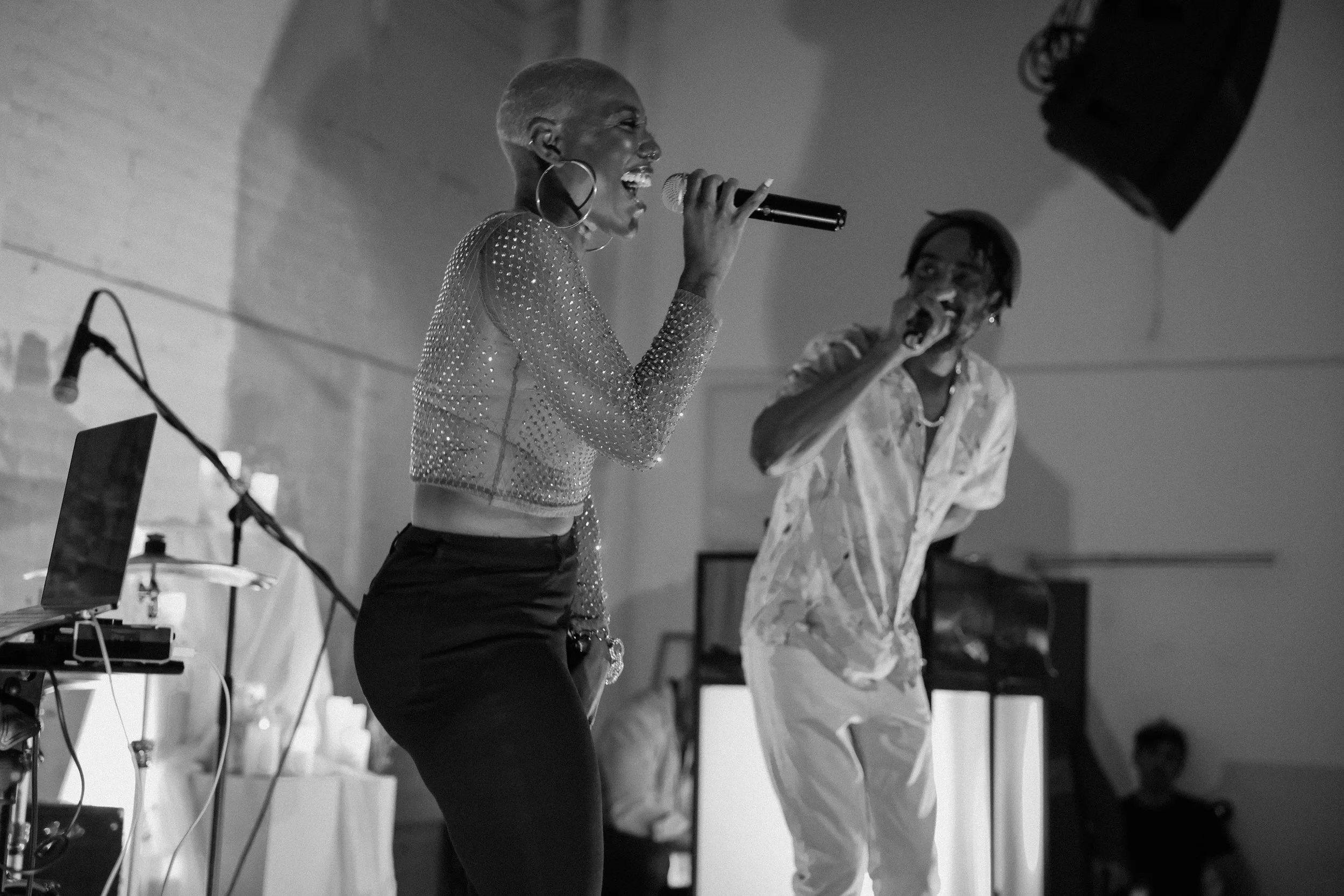 A woman and a man are singing into microphones on a stage, with a DJ setup and speakers visible in the background.