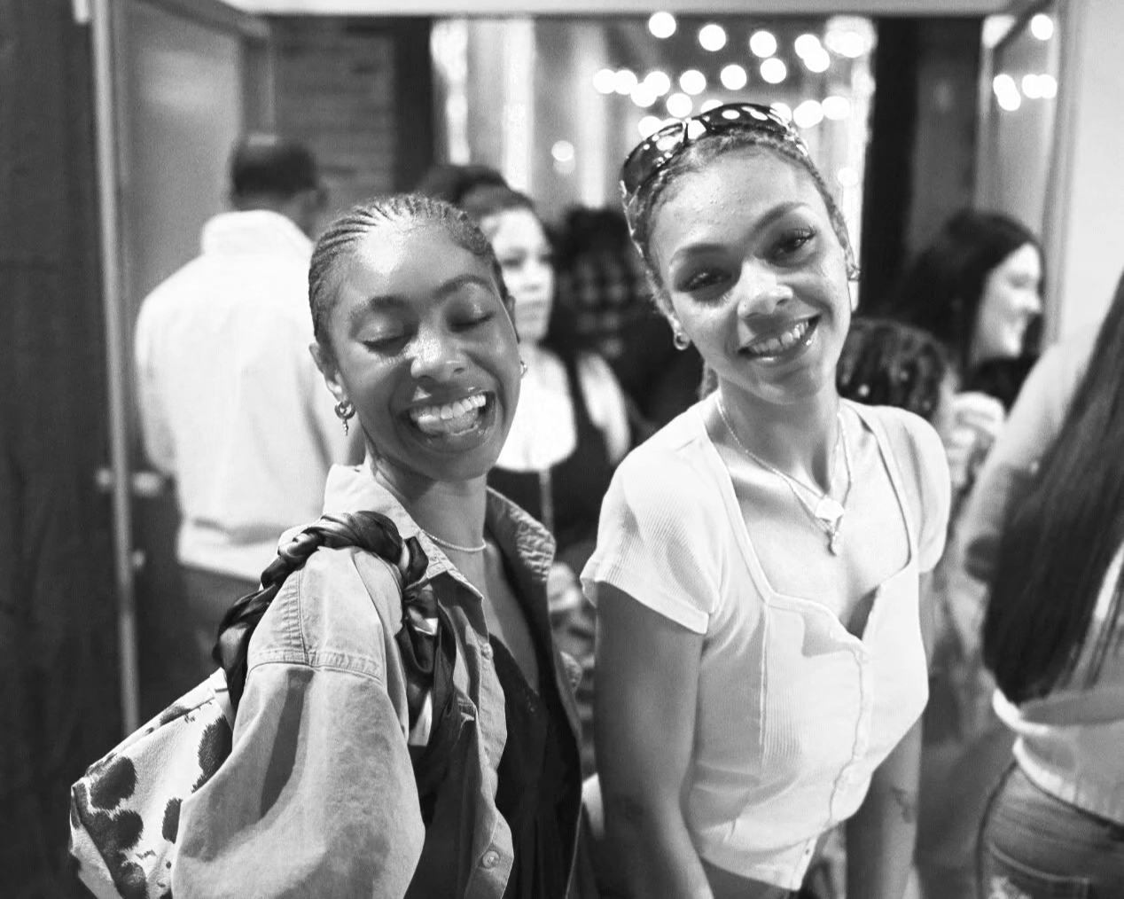 Two smiling women standing close together in a crowded indoor setting, with people in the background, in black and white.