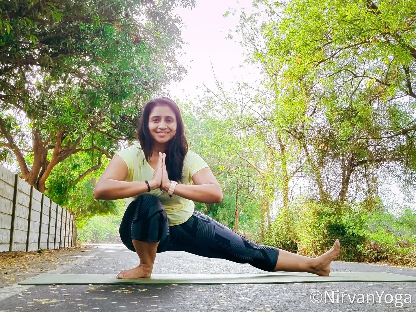 Woman practicing yoga outdoors on a mat, surrounded by trees, performing a side lunge pose with hands in a prayer position, smiling.