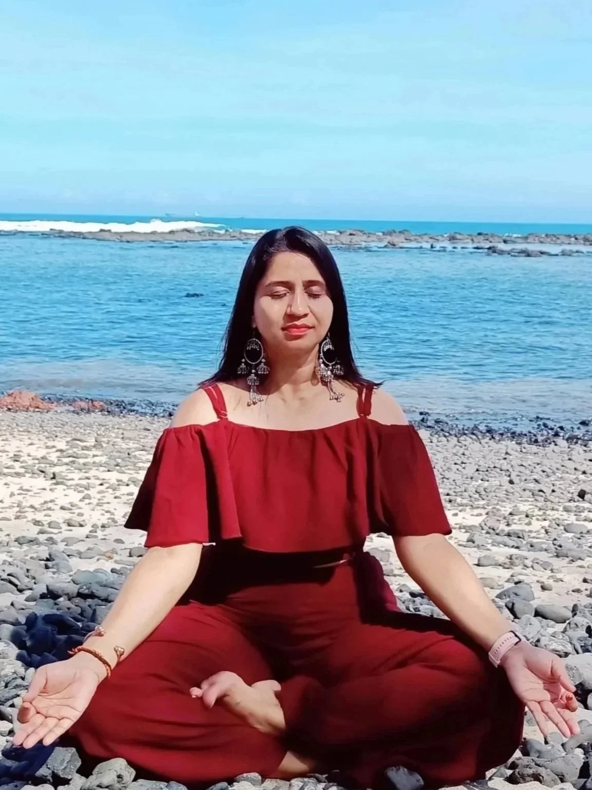 Woman in red clothing practicing meditation on a rocky beach with ocean in the background.