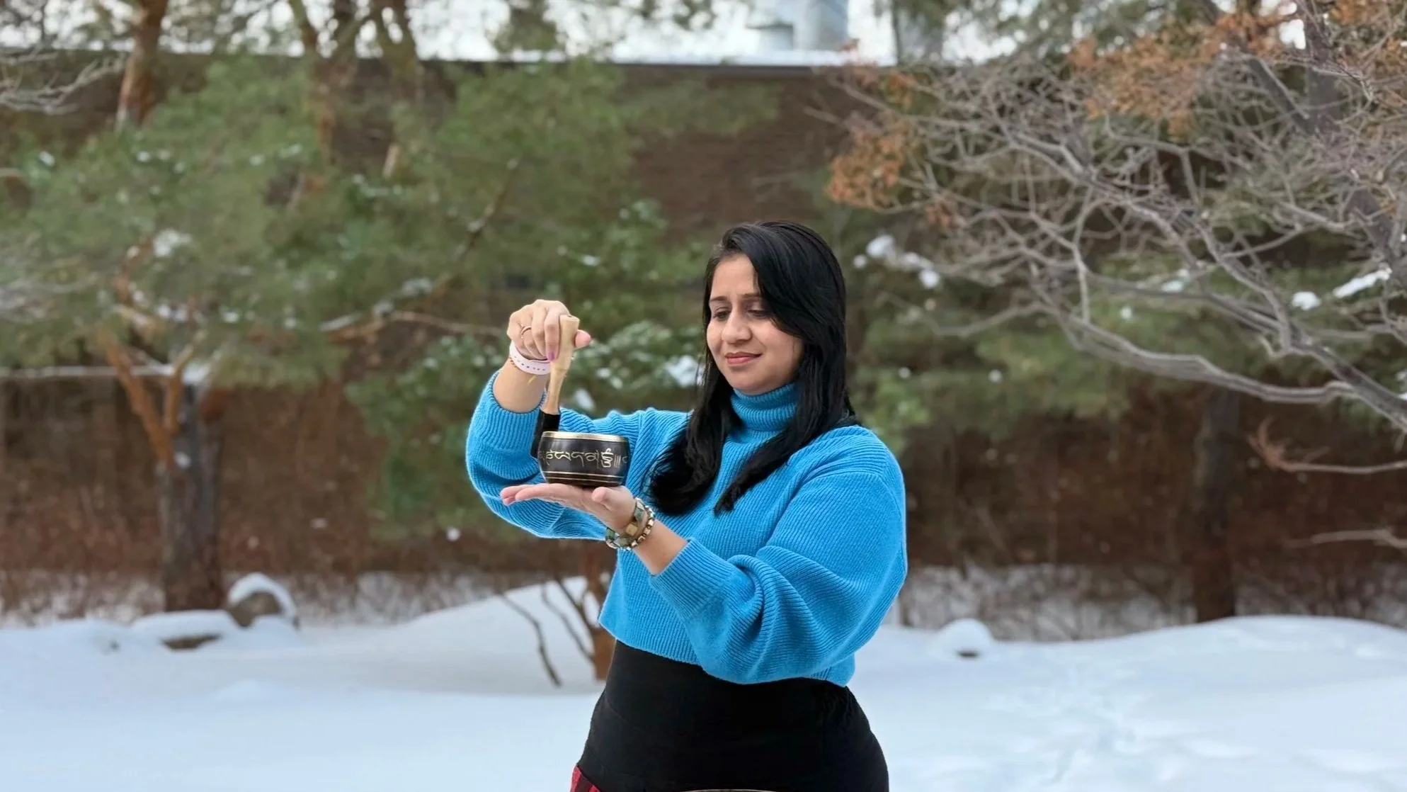 A woman with black hair wearing a blue sweater stands outdoors in a snowy landscape, holding a singing bowl and a wooden striker, with a calm expression.