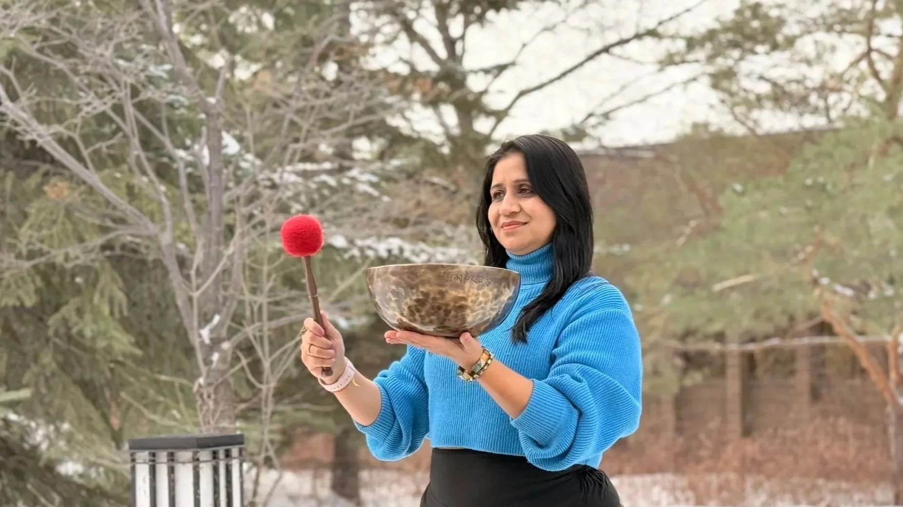 A woman in a blue sweater holding a Tibetan singing bowl in her left hand and a mallet with a red felt tip in her right hand outdoors during winter.