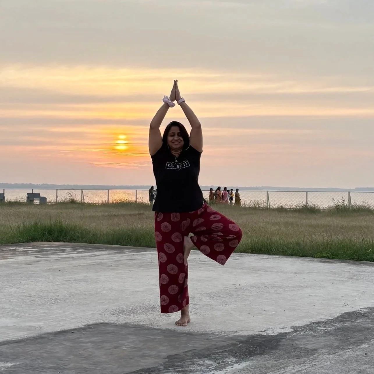 A woman practicing yoga outdoors at sunset, standing on one leg with hands in a prayer position above her head, near a body of water and a fence with a group of people in the background.