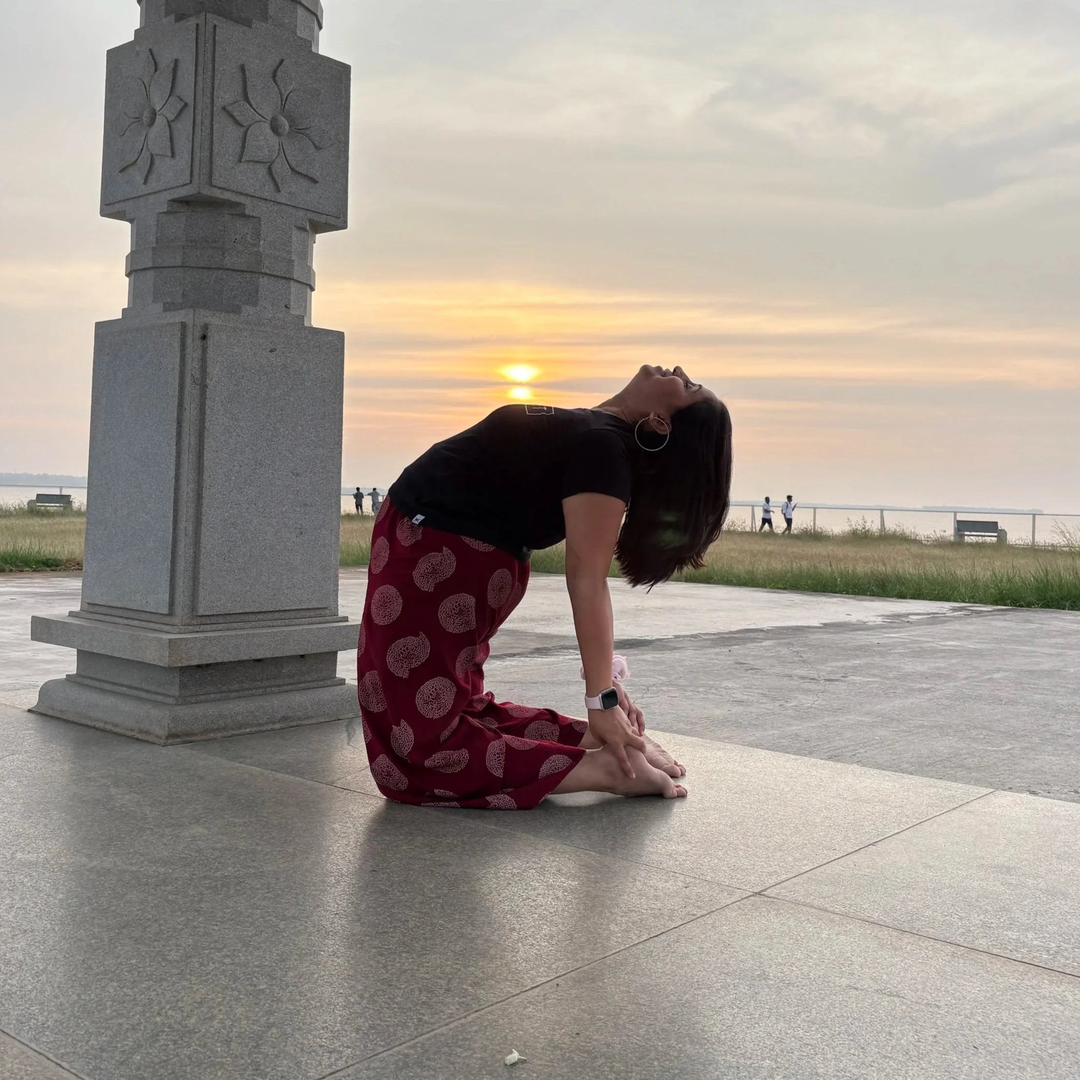 A woman practicing yoga outdoors during sunset, kneeling on the ground with her body arched backward, head tilted up, near a stone monument, with a beach and people walking in the background.