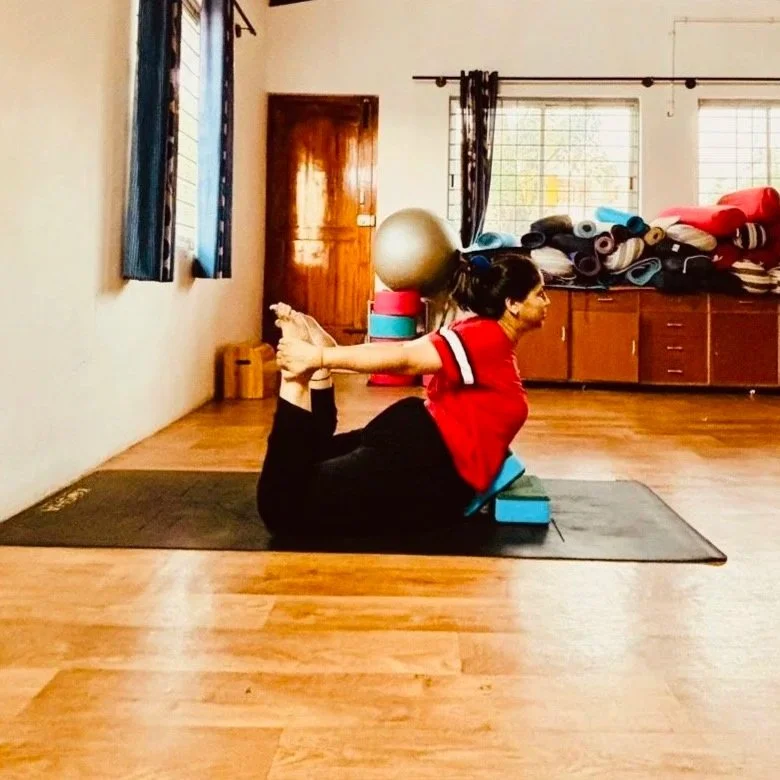 A woman practicing yoga in a studio, lying on a black mat with her legs bent back, holding her ankles, and extending her chest forward with her head turned to the side.