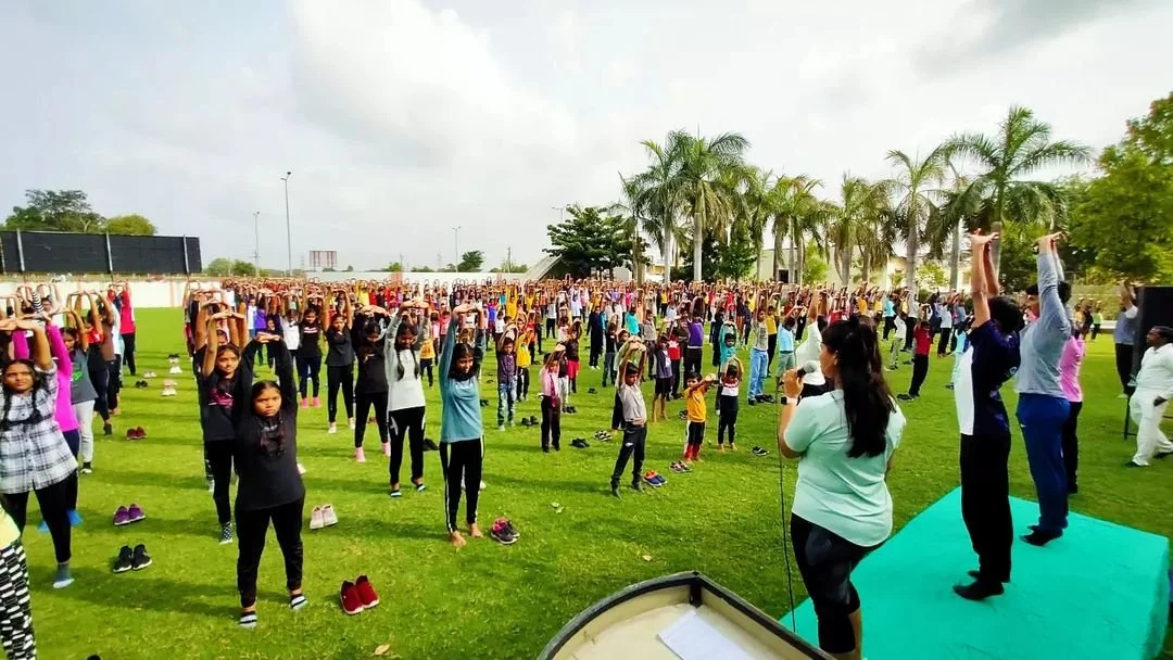 Large group of children and adults practicing yoga outdoors on a grassy field, facing the instructor who is speaking into a microphone, with palm trees and a cloudy sky in the background.