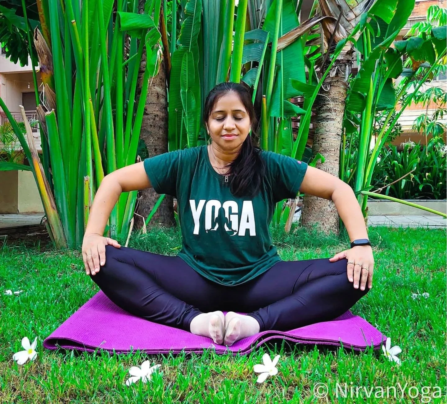 A woman practicing yoga outdoors on a purple mat, sitting in a cross-legged position with hands on her knees, eyes closed, wearing a green T-shirt with 'YOGA' printed on it, black leggings, and a smartwatch, surrounded by green plants and flowers.