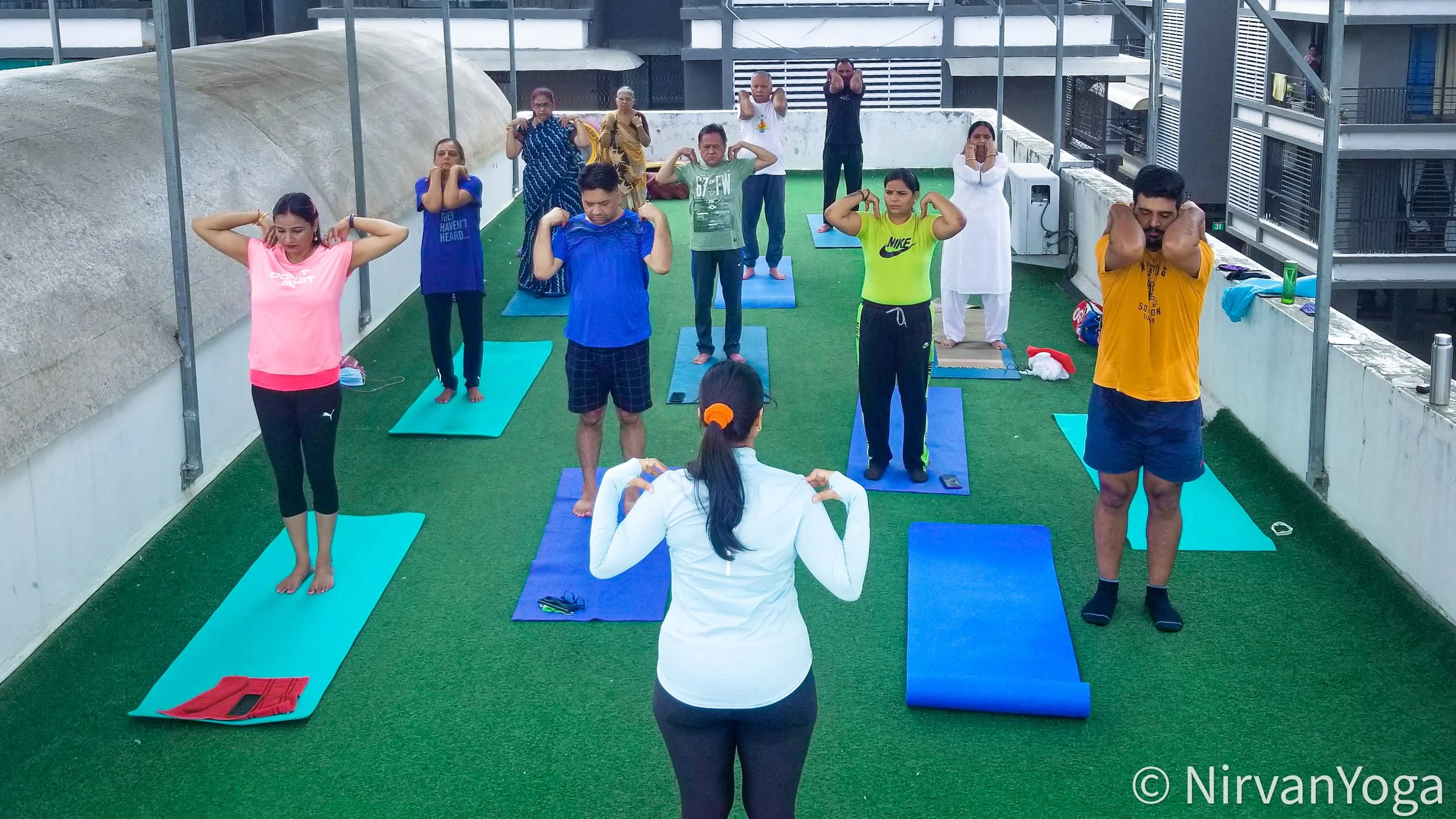 Group of people doing yoga on rooftop terrace with artificial grass, participating in a yoga class led by instructor. Participants are standing on yoga mats, some stretching and others in meditation poses.