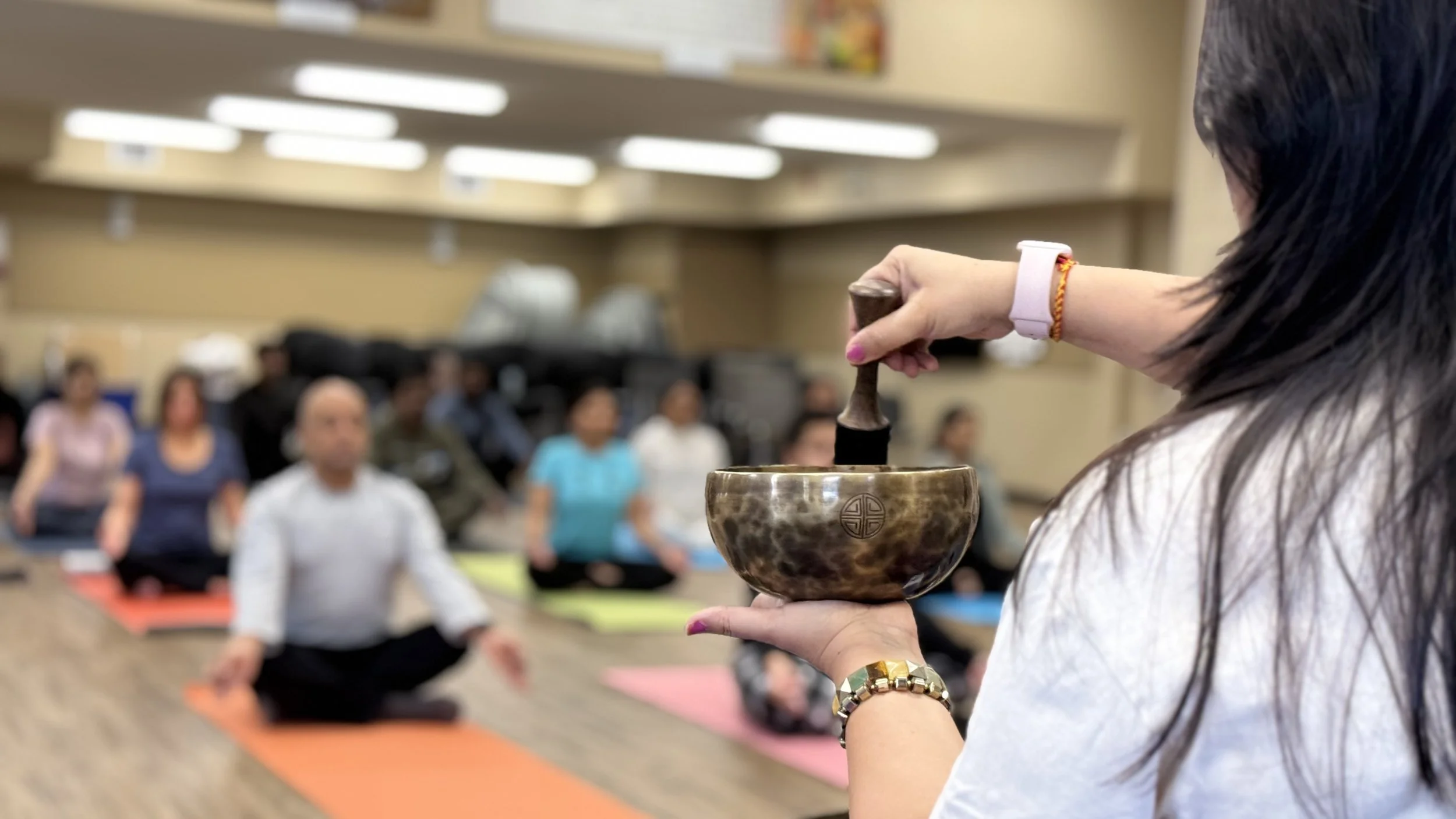A woman in the foreground holding a Tibetan singing bowl with a mallet, leading a yoga or meditation class with participants seated on colorful mats in a spacious room.