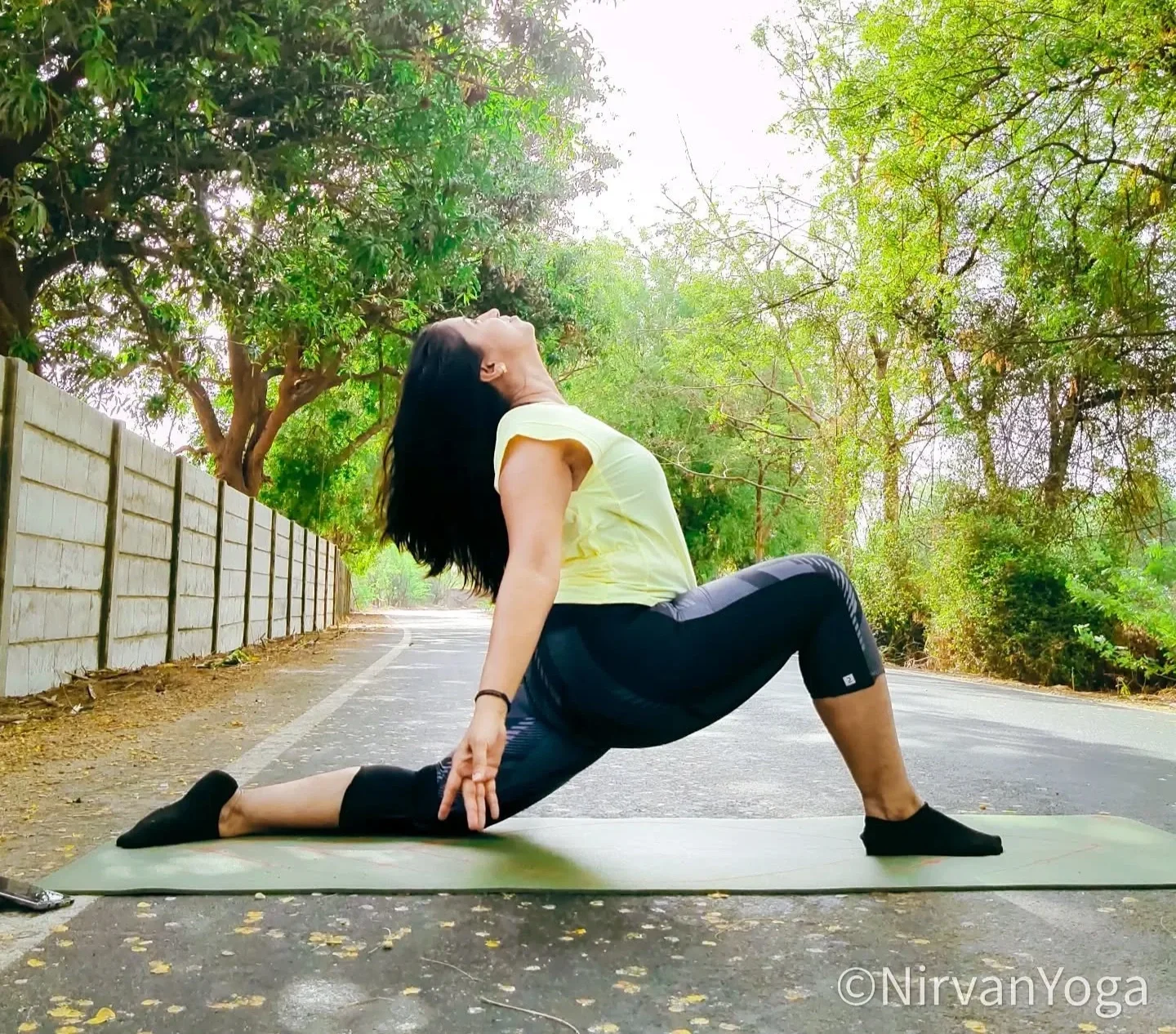A woman in a yellow shirt and black leggings practicing yoga outdoors on a green mat on a paved road, performing a low lunge pose with her head tilted back and arms extended behind her.