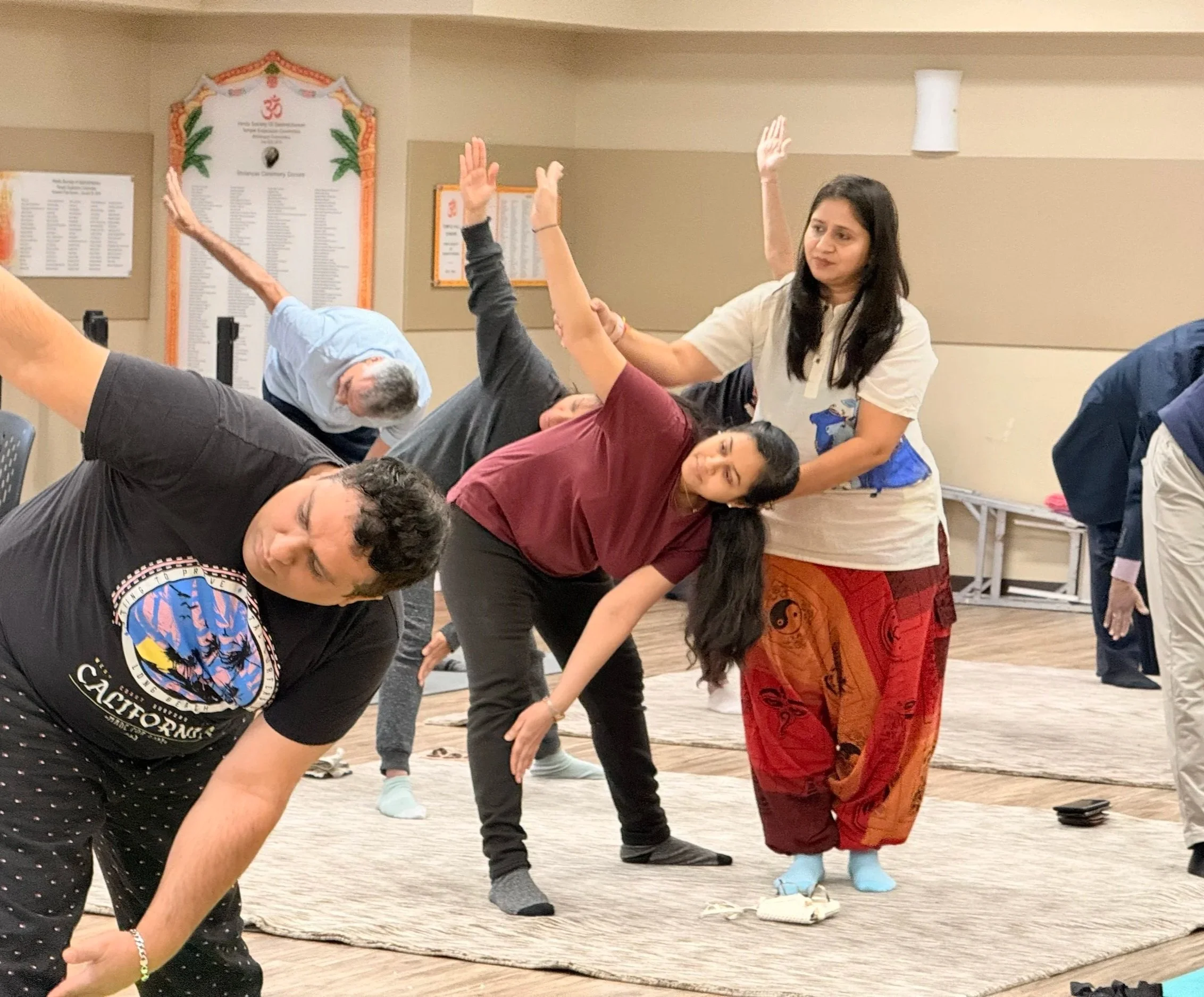 Group of people practicing yoga on mats indoors, with a woman assisting a participant in stretching.