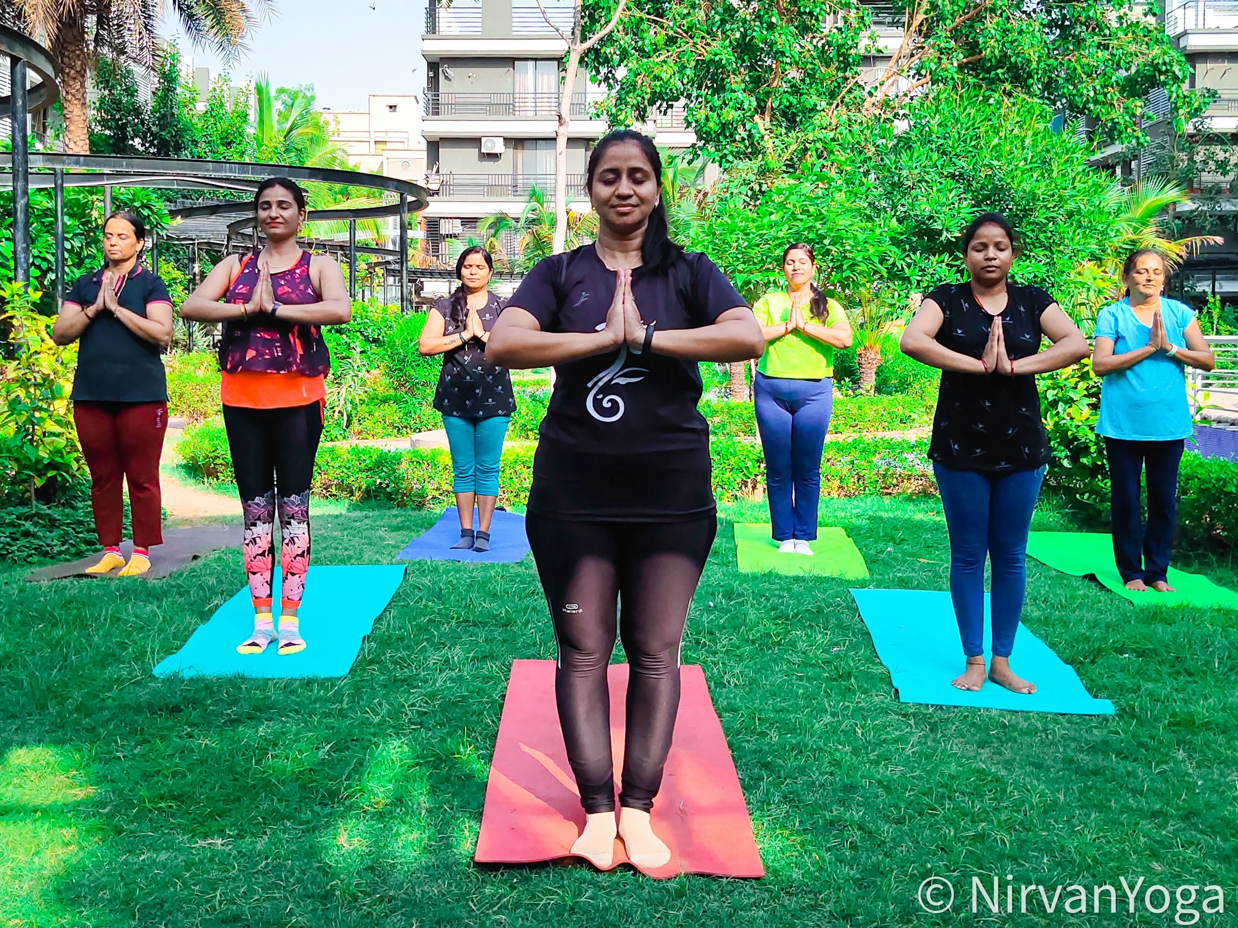 Group of women practicing yoga outdoors on colorful mats in a lush garden with trees and buildings in the background.