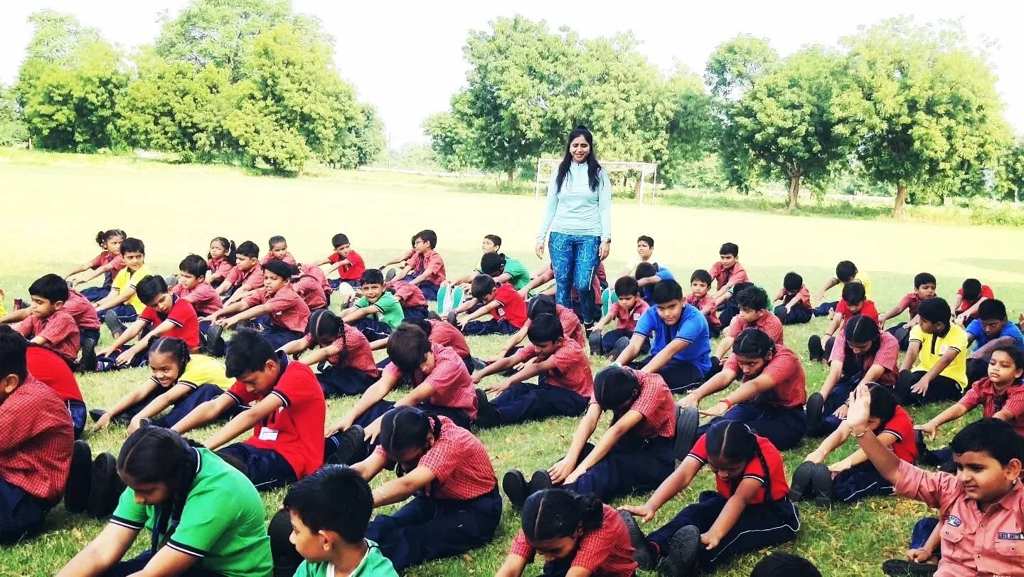 Group of school children sitting on grass doing stretching exercises outdoors, with a female teacher supervising.
