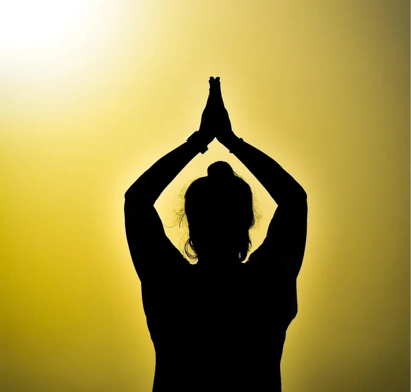 Silhouette of a person with hair up, hands in prayer position above head, against yellow background.