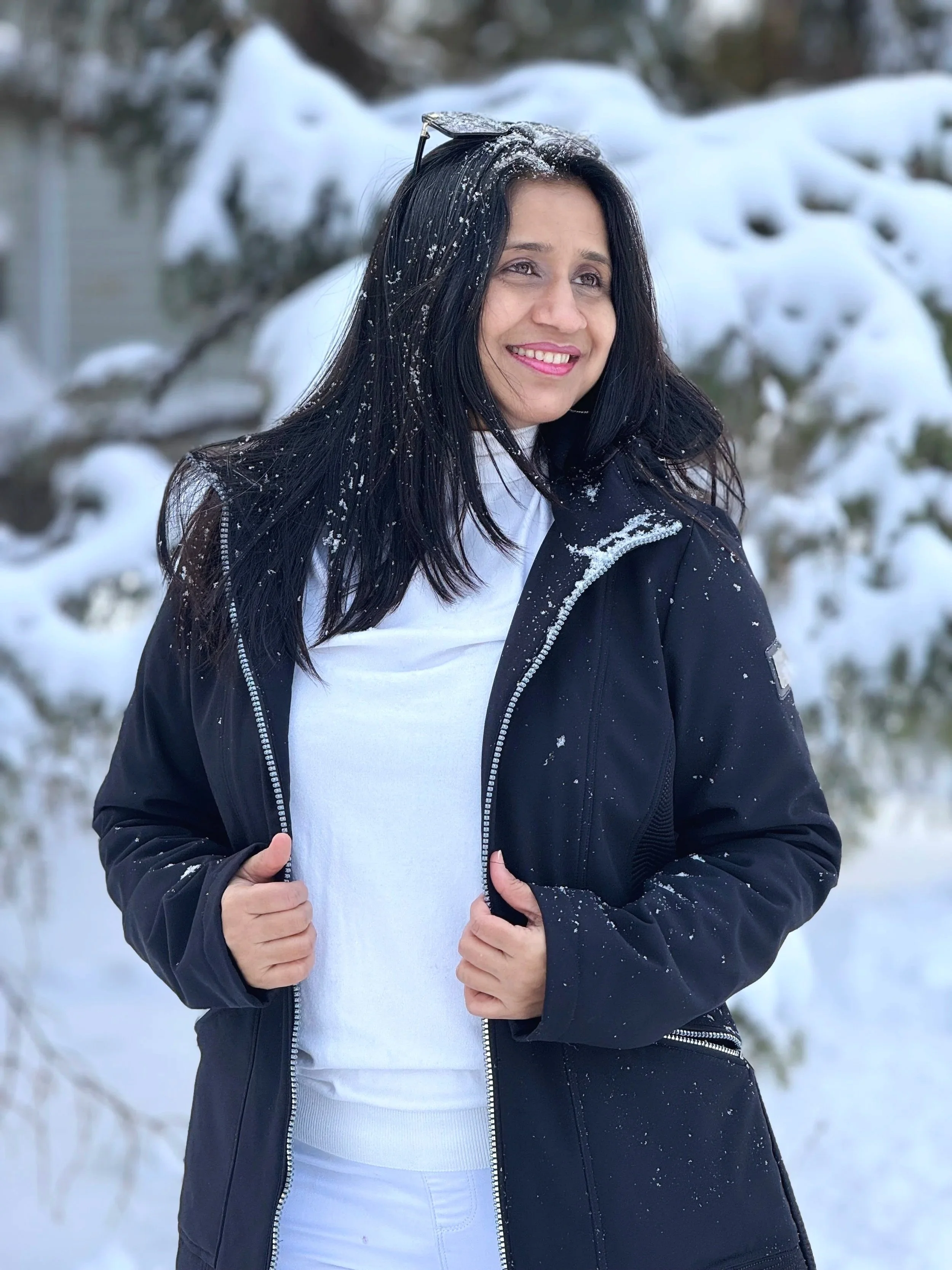 Woman with black hair smiling outdoors in snowy landscape, wearing a black jacket and sunglasses on her head.