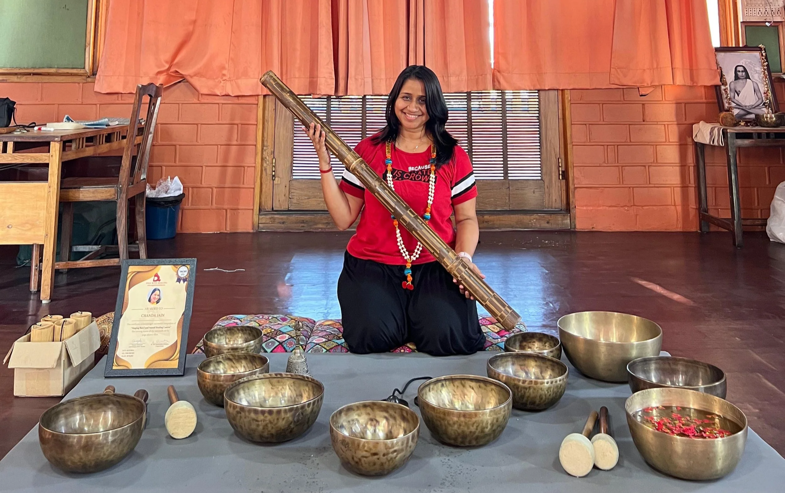 Woman sitting on the floor holding a long musical instrument, surrounded by singing bowls and tuning mallets, in a room with orange curtains and wooden furniture.