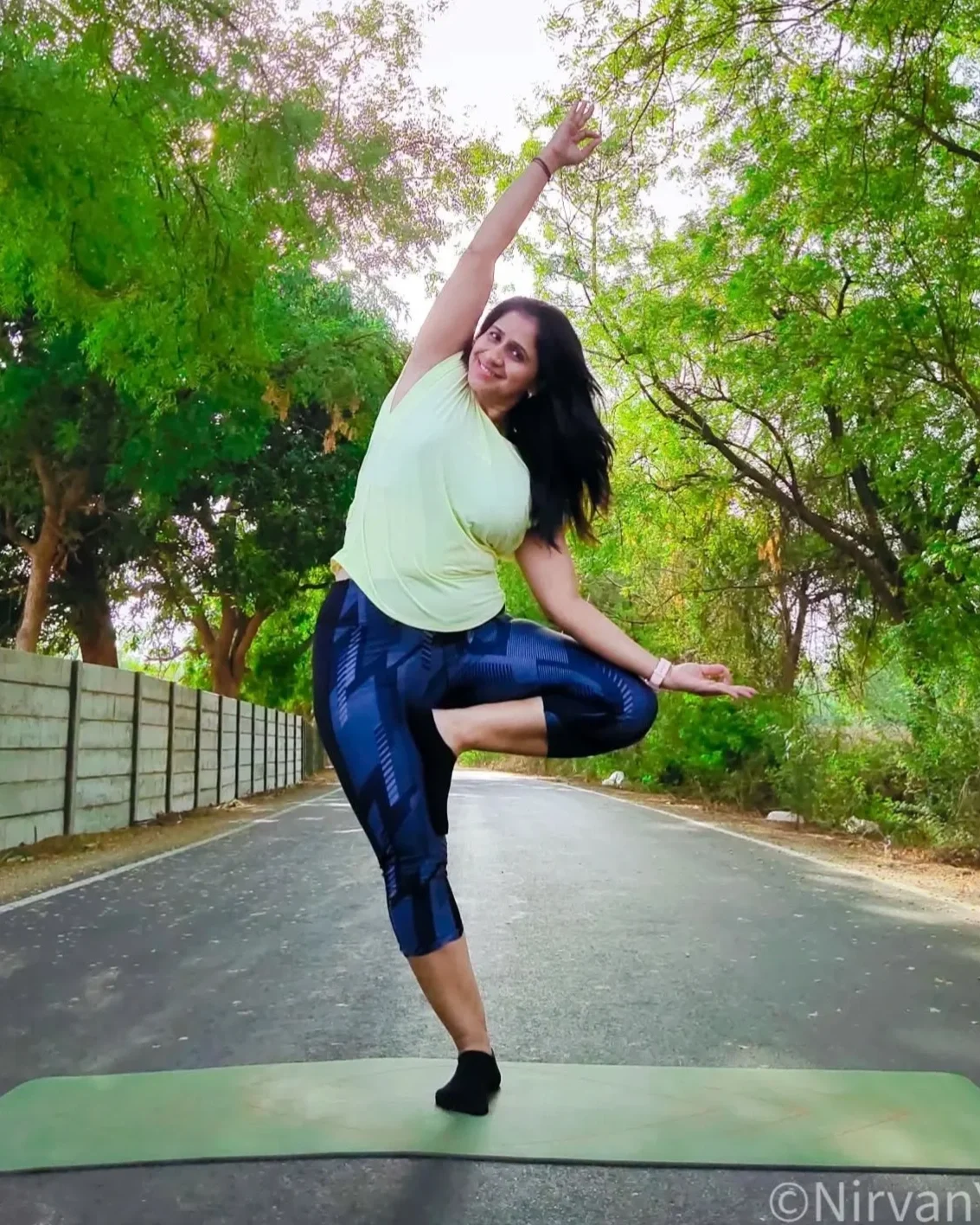 A woman doing a yoga pose on a green yoga mat on an empty road surrounded by green trees.