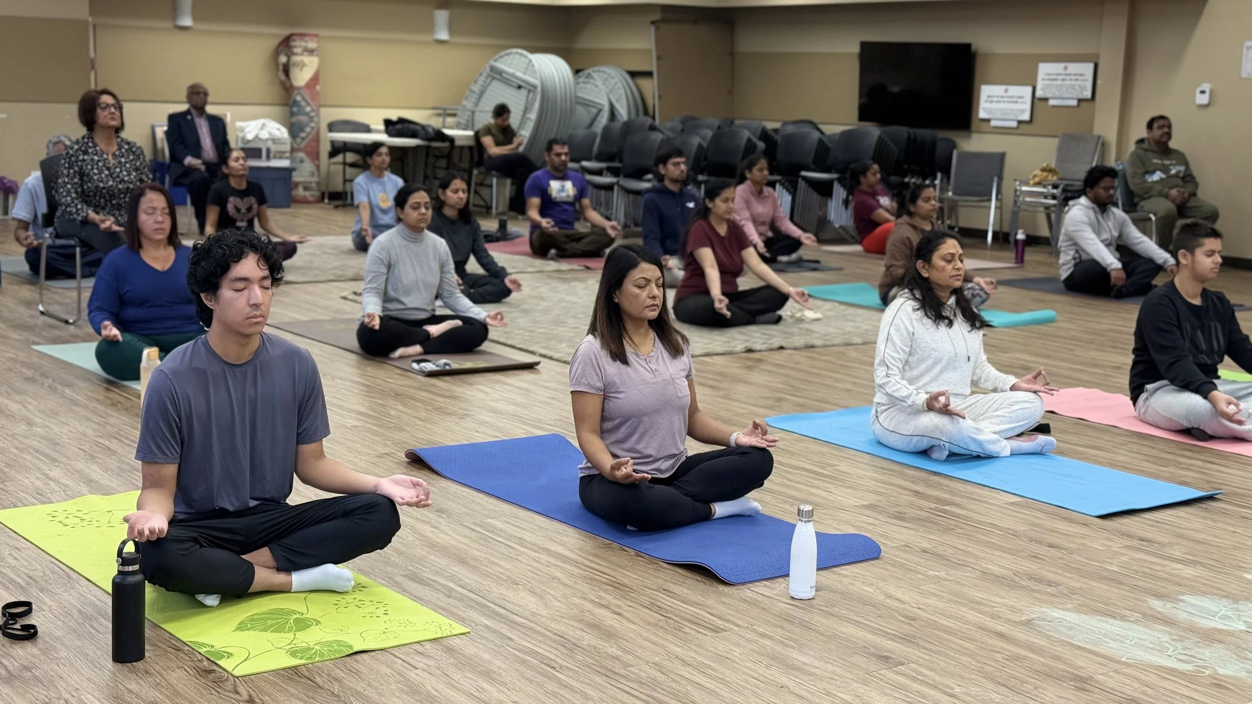 Group of people meditating in a yoga class, seated on mats in a spacious room with wooden floors.