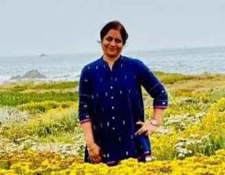 Woman in a blue traditional outfit smiling outdoors near a grassy shore with the ocean in the background.