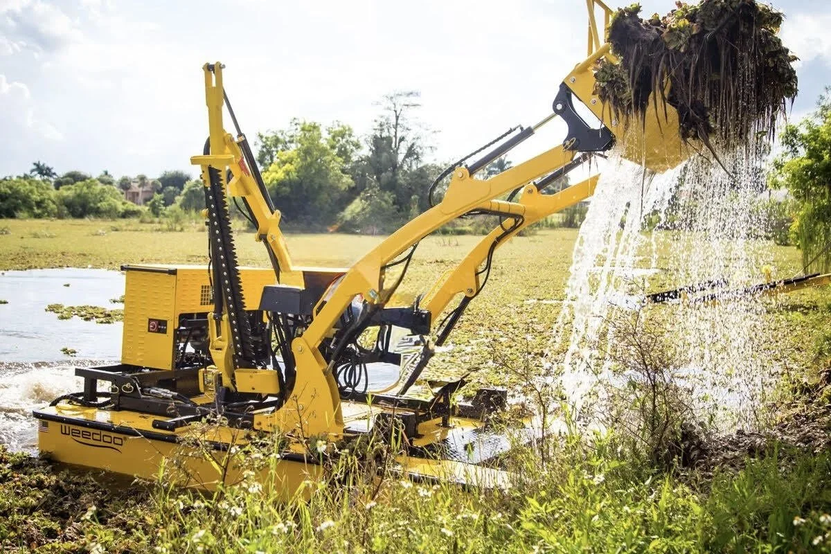 A "Weedoo" boat removing debris and weeds from the water's edge near a lake, with a grassy shoreline and trees in the background.