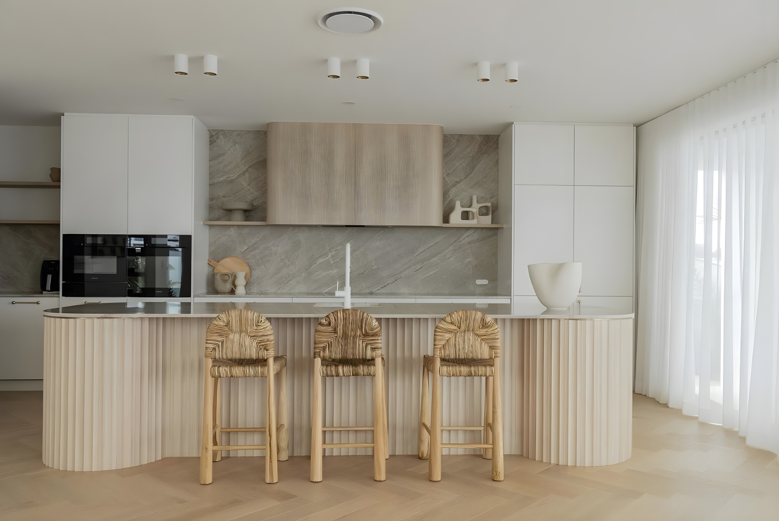 Luxury kitchen with curved island, woven stools and soft neutral finishes.
