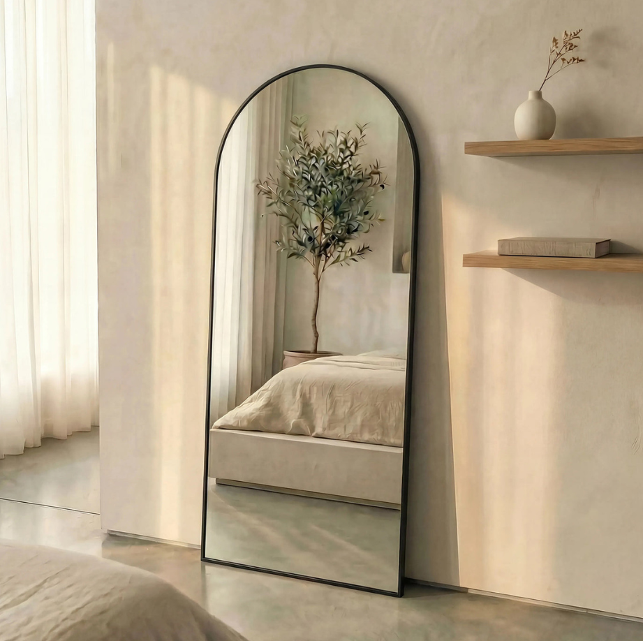 Arched floor mirror styled in warm bedroom reflecting layered bedding and indoor greenery.