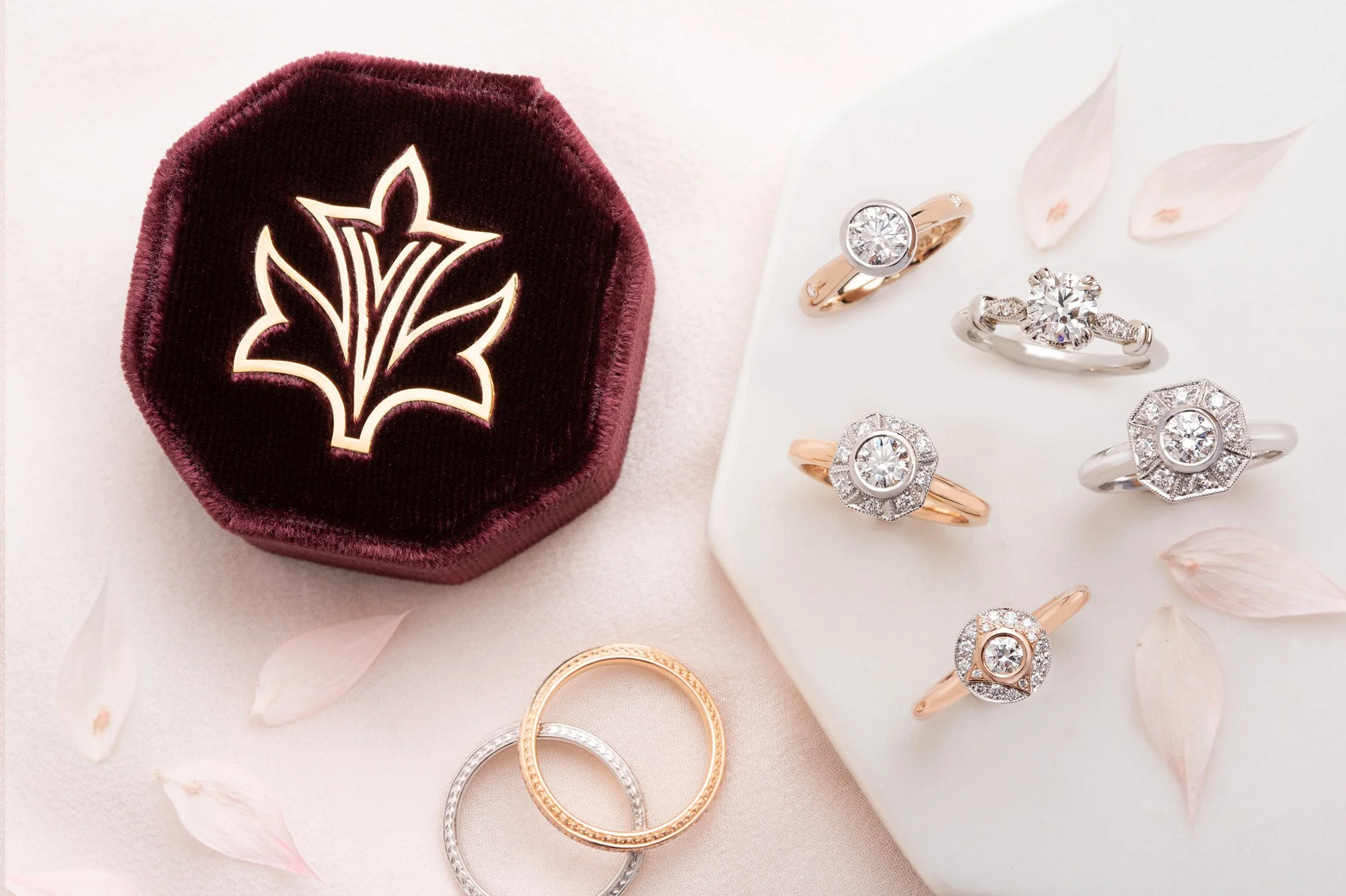 Jewelry display with diamond rings on a white marble slab, pink flower petals, and a velvet jewelry box.