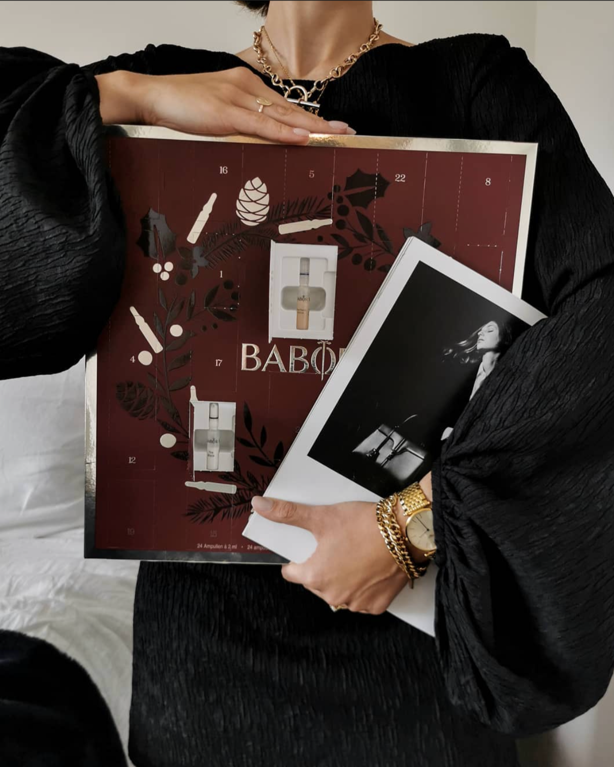 Person wearing a black dress with textured fabric, gold jewelry, holding an Advent calendar and a black-and-white photograph, standing in an indoor setting.