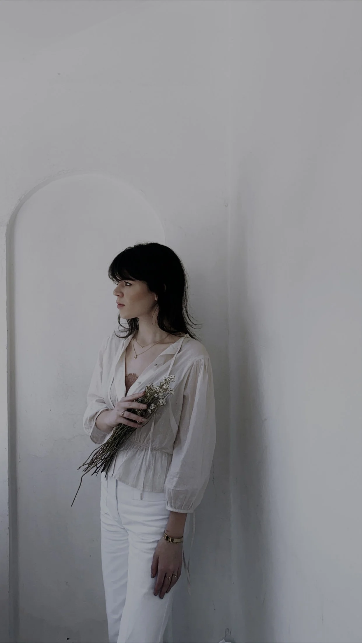 A woman with dark hair holding a small bouquet of wildflowers, standing against a plain white wall, wearing a cream-colored blouse and white pants, looking to the left.