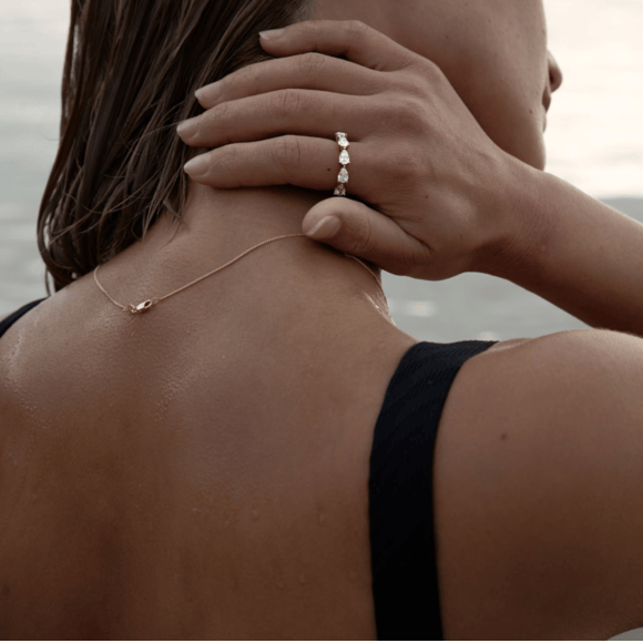 Close-up of a woman touching the back of her neck, showing a diamond ring on her finger and a delicate necklace, with a blurred outdoors background.
