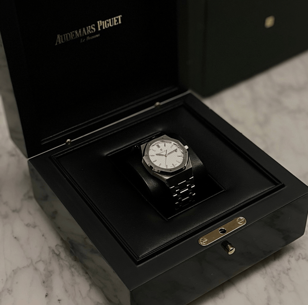 A luxury wristwatch with a white face and metal band inside an open black display box on a marble surface.