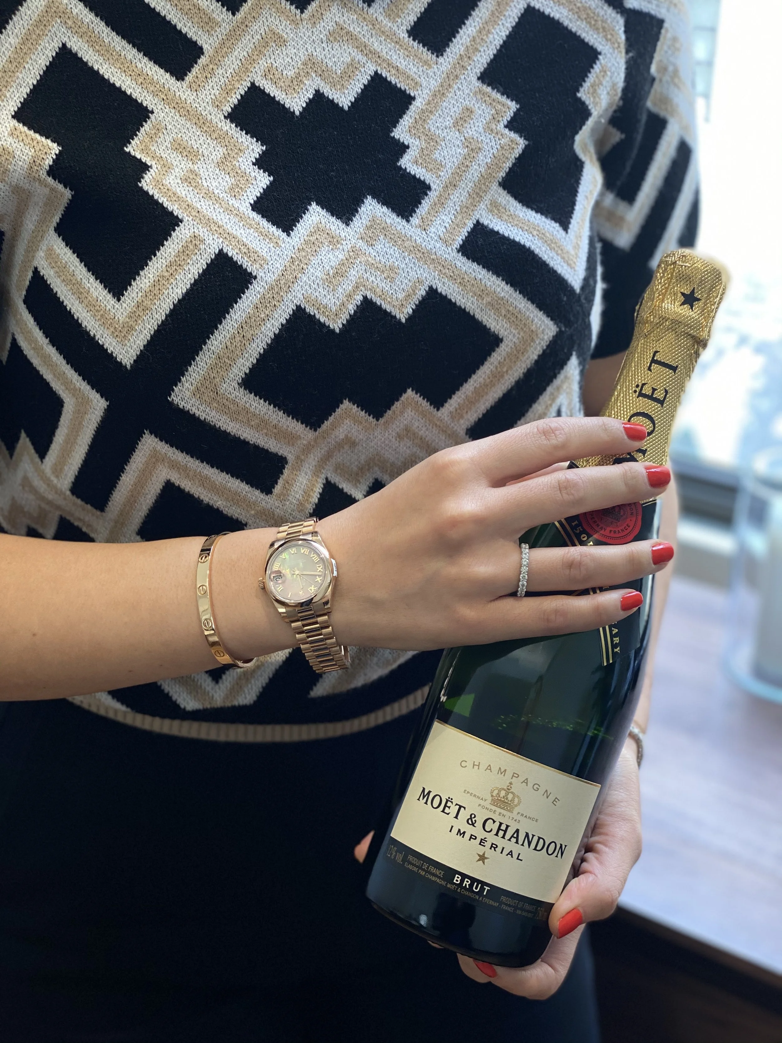 A woman holding a bottle of Moët & Chandon champagne, wearing a patterned black, beige, and white top, a gold watch, a gold bracelet, and a diamond ring, with red nail polish.