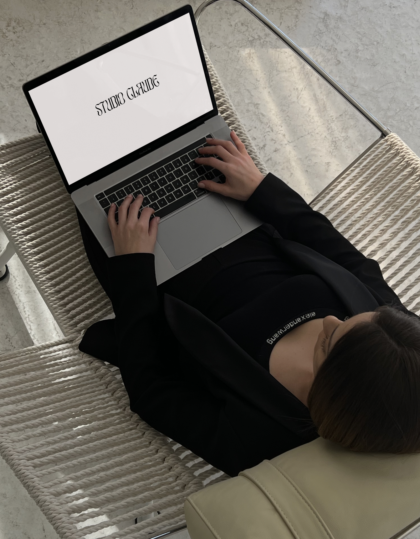A woman lying on a striped bench, working on a laptop with 'STUDIO GLAM' displayed on the screen. She is wearing a black outfit and has a beige bag beside her.