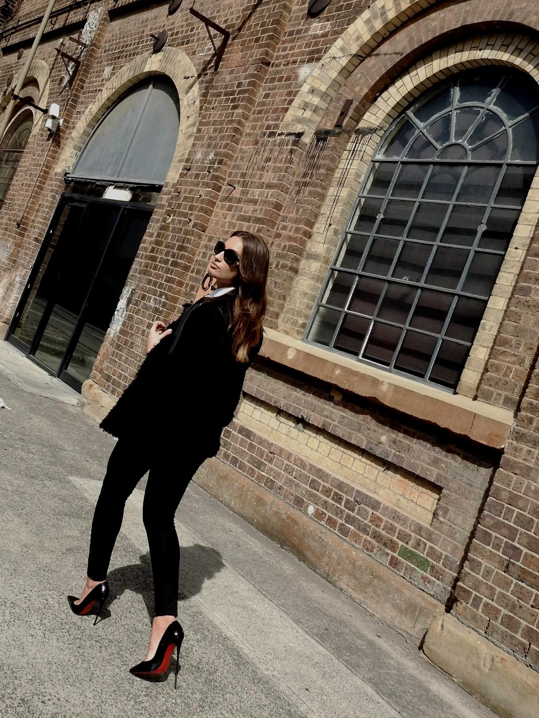 A woman dressed in black with high heels, sunglasses, and earrings, standing against a brick building with arched windows, on a sidewalk in bright sunlight.