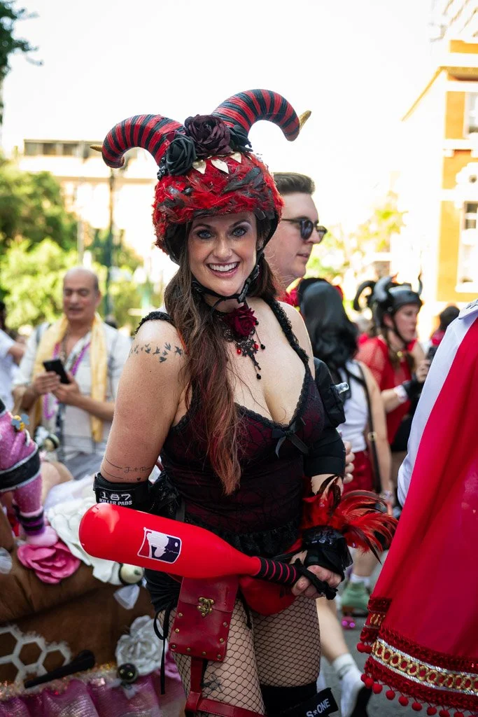 Woman dressed in a devil costume with red and black horns, red wig, fishnet stockings, and holding a red and black prop bat, standing outdoors among people at a street event.