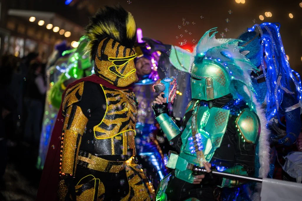 Two people in elaborate, brightly colored costumes with neon lights and LED decorations, engaging in a street festival or parade at night.